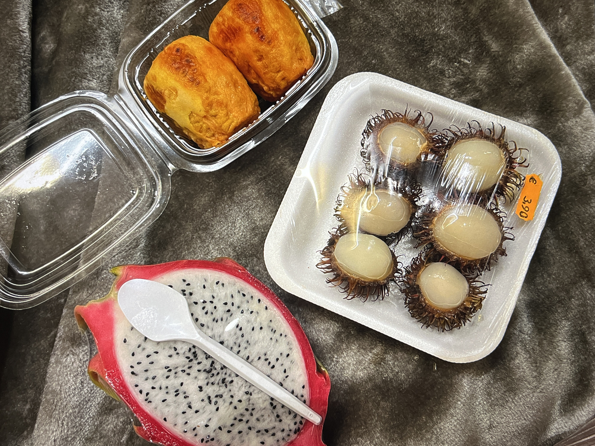 The image shows a still life of food items arranged on a gray, textured surface, possibly fabric. On the left, there's a clear plastic container holding two baked goods, golden-brown in color. Below that, a halved dragon fruit is visible, its vibrant pink rind contrasting with the white flesh dotted with black seeds. A white plastic spoon rests on the dragon fruit. To the right, a white foam tray contains several rambutans, each with a translucent white fruit partially visible through its spiky, brown outer layer. A price tag reading "€ 3.90" is affixed to the tray. The location appears to be Unknown, Unknown.