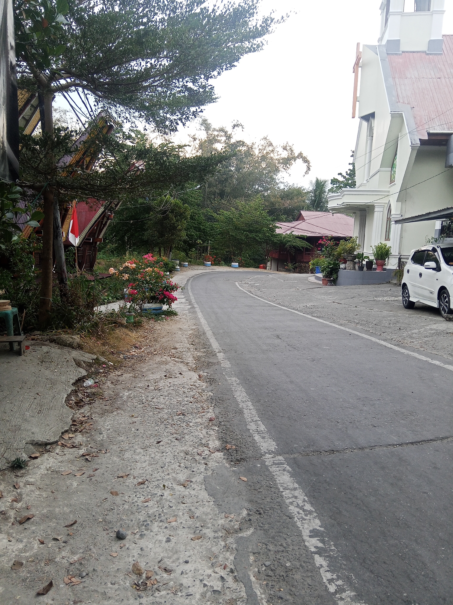The image depicts a street scene in Makale, Indonesia, with a focus on a curved asphalt road. On the left side of the road, a concrete walkway is visible, bordering a lush area with a tree, some flowering bushes with pink and orange blooms, and a traditional-looking wooden structure adorned with a red and white flag. Further down the street on the right, a white car is parked in front of a modern white church with a red roof. The environment is verdant, with trees and foliage present throughout the scene. The sky is overcast, suggesting a cloudy day. The road has a white dashed line marking the lane division. The ground is uneven in places, with scattered leaves and debris.