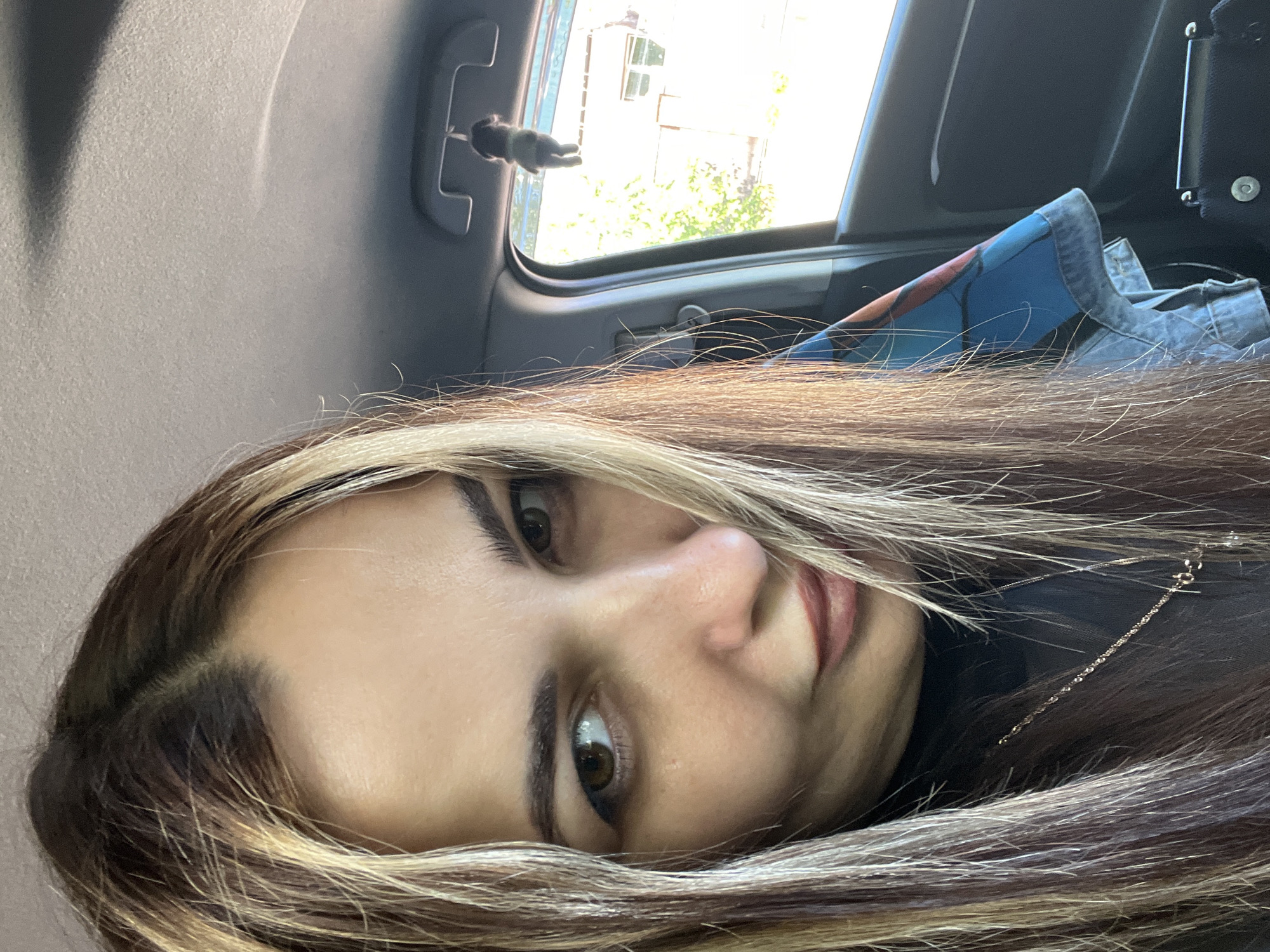 A close-up image shows a woman with brown and blonde highlighted hair, partially obscuring her face. She appears to be inside a vehicle. The interior of the vehicle shows a light gray ceiling, a window with a toy figure hanging from the handle, and a blue and denim object. The location is Unknown, Unknown.