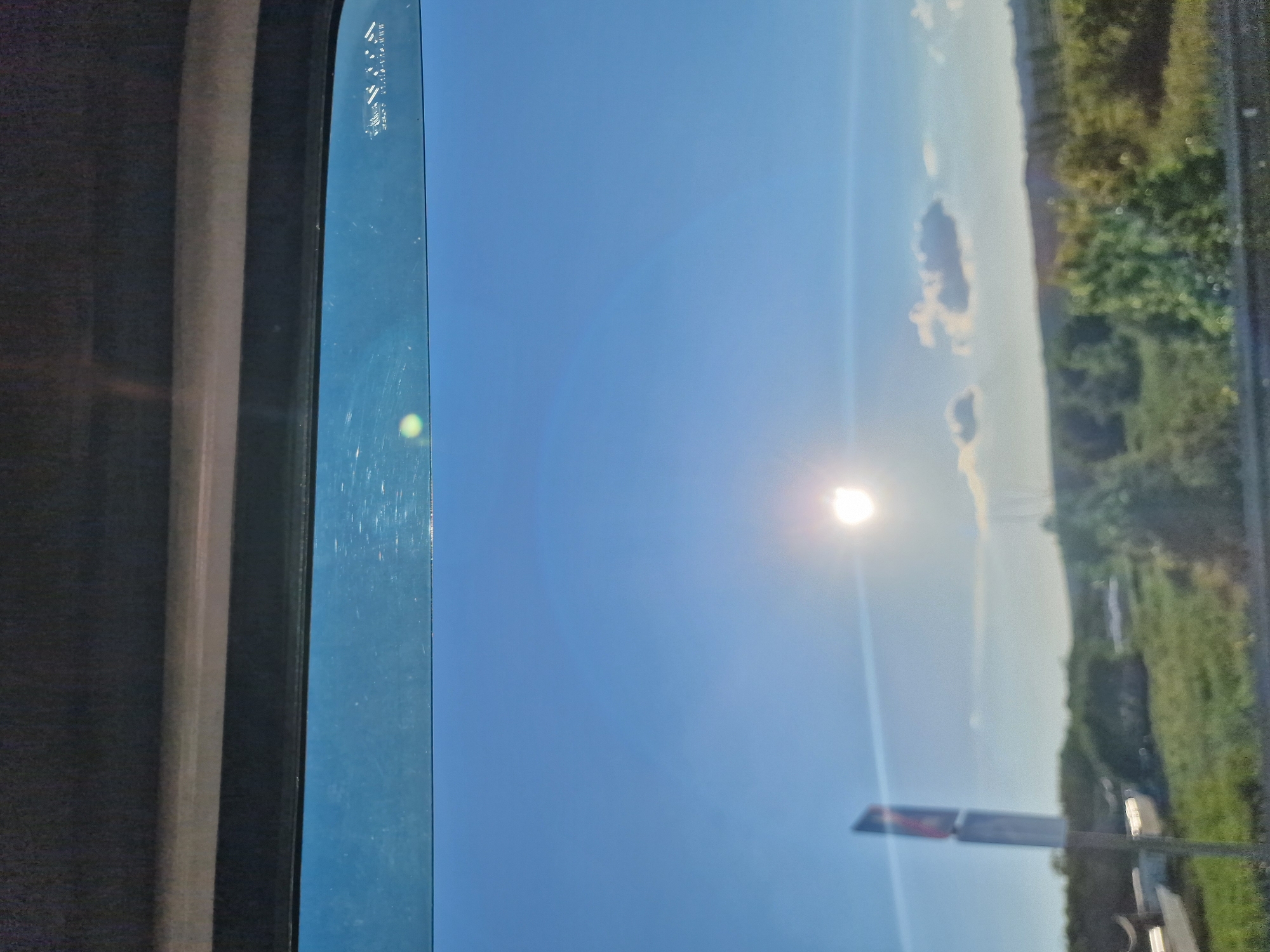 The image captures a bright, sunny daytime scene viewed from inside a vehicle, looking out through a window. The perspective is rotated 90 degrees clockwise from standard orientation. A vast, clear blue sky dominates the view, with the sun positioned centrally, creating a prominent lens flare. A few small, scattered white clouds are visible in the distance.
On the left, the dark interior frame of the vehicle's window is visible, with the blue-tinted glass showing numerous scratches and a small, greenish-yellow circular reflection or flare. Faint, white text, likely manufacturing details or a vehicle identification number, is etched into the upper left corner of the window glass; it is mostly illegible, but fragments like "TOP AS M" and other characters are discernible.
The lower portion of the scene depicts a landscape with a hazy horizon line, suggesting distance. Green trees and bushes line what appears to be the edge of a road or embankment on the right side. A partially visible road sign, with dark lettering on a lighter background, stands upright in the lower right quadrant. The overall impression is of a clear and pleasant day, likely while traveling in Tatarka, Russia. No people are visible in the image.