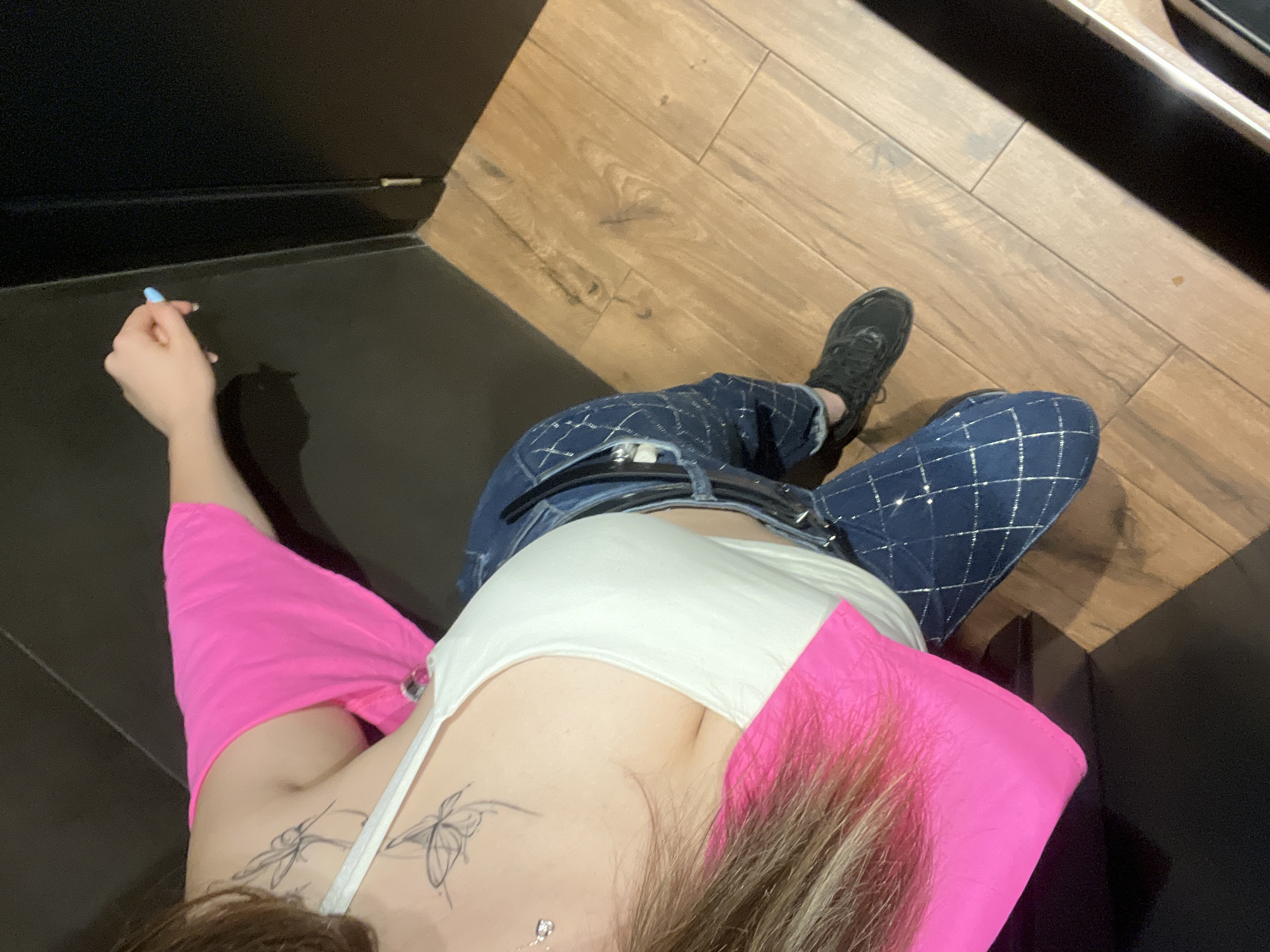 The image shows a woman in a white tank top and bright pink short-sleeved top, wearing blue jeans with a diamond pattern and black sneakers. She has tattoos of butterflies on her upper arm and shoulder. She is holding a light blue object, possibly a vape pen, in her right hand. The woman is standing on a wooden floor next to a black wall, with a shadowed area to her left and a darker area with a vertical seam to her right. The lighting suggests an indoor environment, potentially a public restroom or a changing area within a venue, due to the dark walls and wooden floor. The overall scene is consistent with a casual indoor setting in Plovdiv, Bulgaria, though specific landmarks or identifying features are absent.