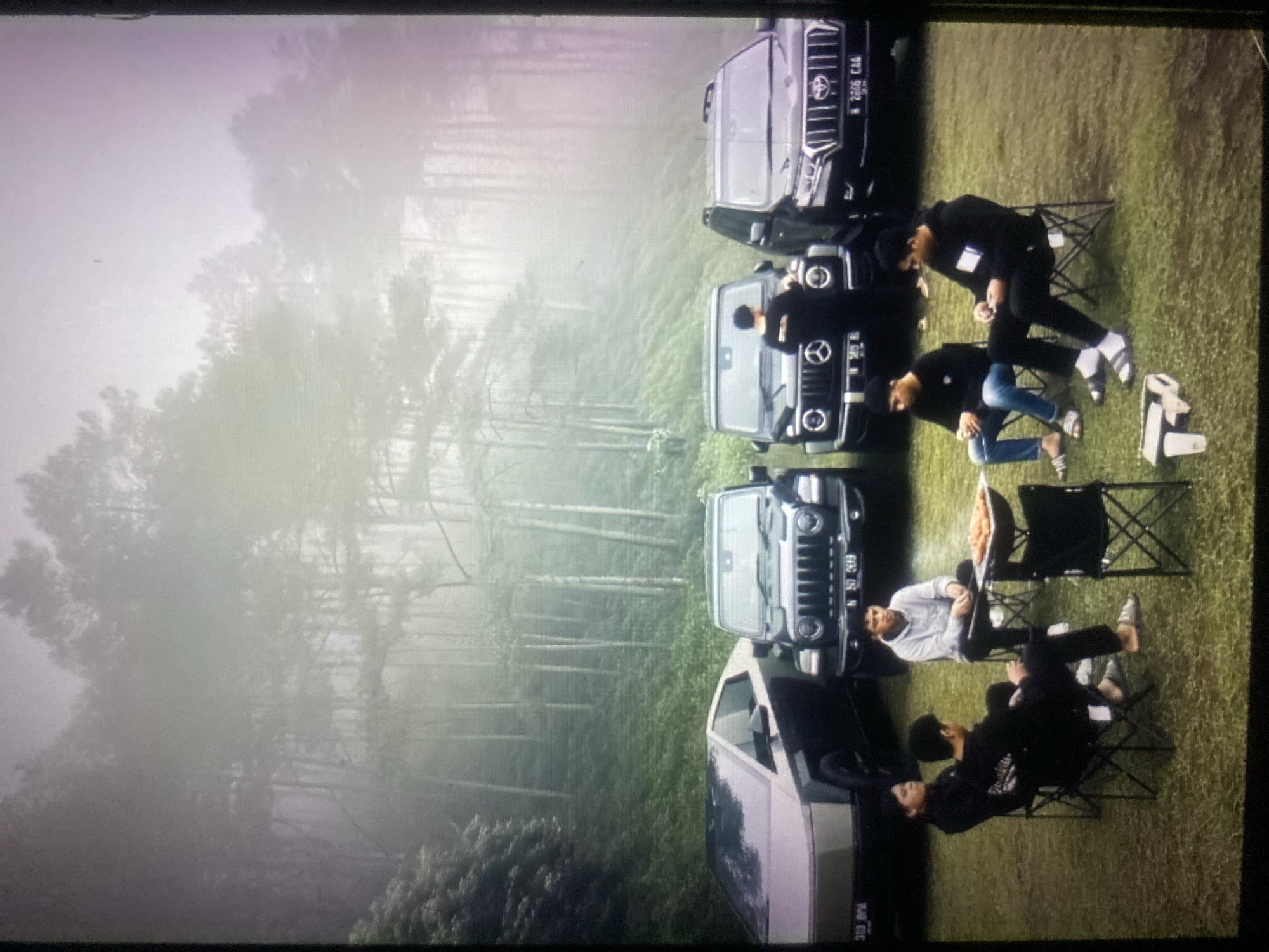 The image depicts a group of six men gathered in a grassy clearing, likely a campsite, with a backdrop of dense, misty forest. Four off-road vehicles are parked in the background: a Tesla Cybertruck on the far left, followed by a black Jeep Wrangler, a black Mercedes-Benz G-Wagen, and a black Toyota Land Cruiser Prado on the far right.
In the foreground, five men are seated on folding chairs. One man, wearing a light grey hoodie, sits facing a small barbecue grill laden with food, appearing to be in the process of cooking. To his left, a man in a blue hoodie and jeans is seated, holding a phone. Further to the left, two men in black long-sleeved shirts are seated next to each other. The leftmost man is looking away from the camera, while the man next to him appears to be engaged with his phone. A sixth man, also in a black long-sleeved shirt, is standing near the Mercedes-Benz G-Wagen. The ground is covered in grass, and the overall atmosphere is subdued, with the fog lending a serene and possibly cool ambiance to the mountainous or hilly terrain.