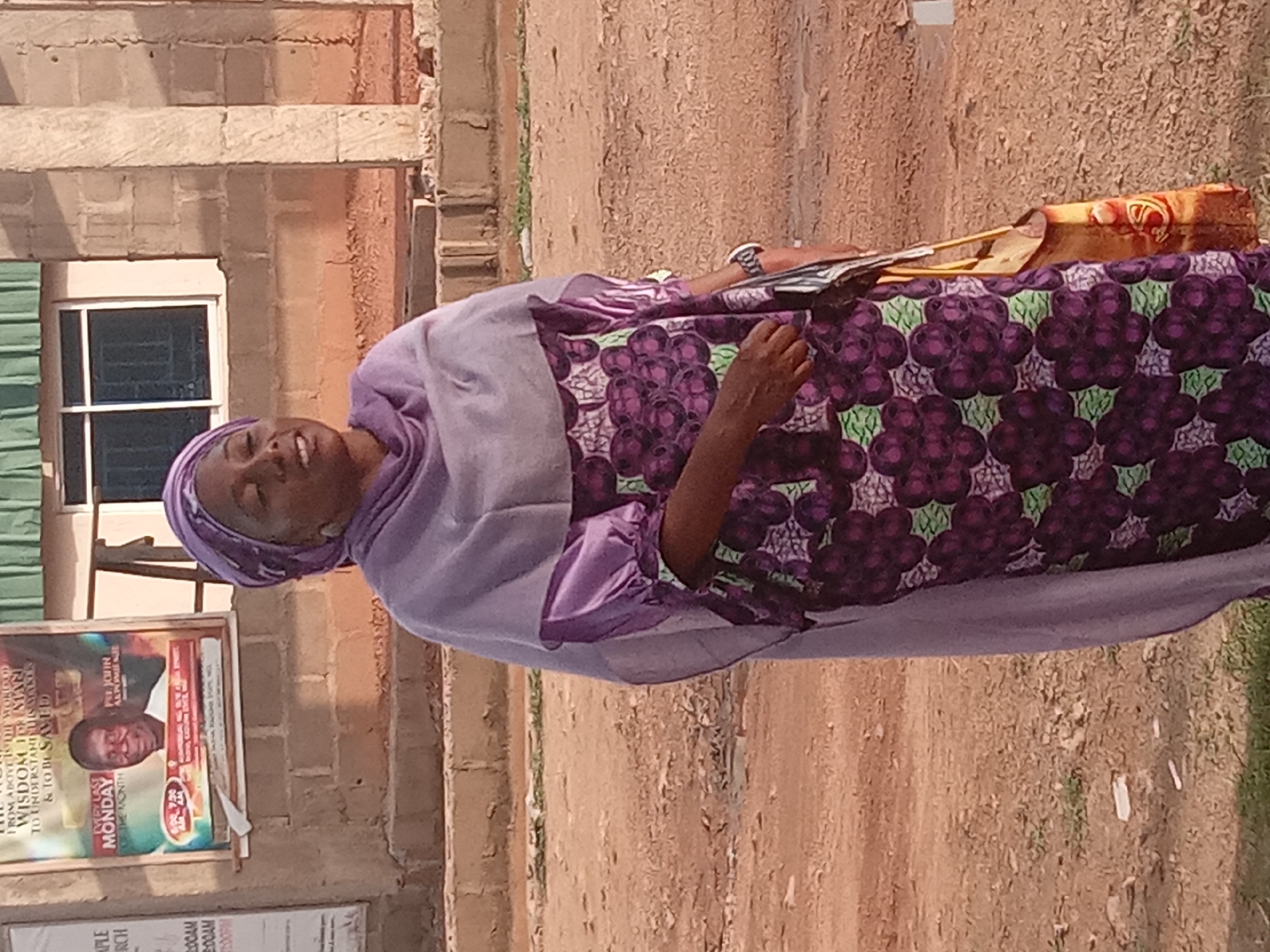 A woman wearing a purple headscarf and a long purple dress with a floral pattern stands outdoors. She is holding a light brown bag in her right hand. Behind her, there is a brick wall and a window with green blinds. A poster with an image of a man and text is visible on the left side of the frame. The ground in front of her is dirt with some patches of grass.