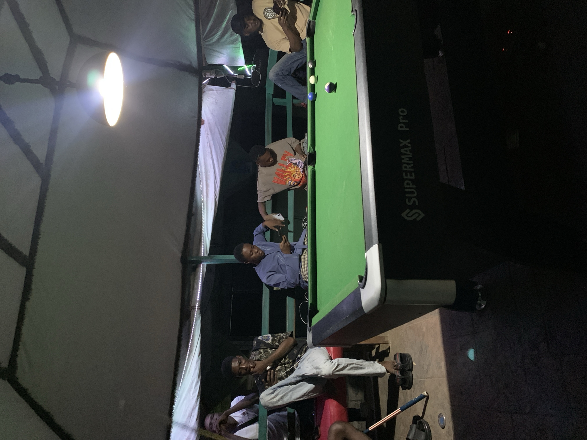 The image depicts a group of young men gathered around a pool table, seemingly at an outdoor establishment. The scene is dimly lit, suggesting it is nighttime or evening. The setting appears to be a covered area, possibly a patio or a covered outdoor space, with a large hanging light fixture providing illumination.
There are at least five individuals visible in the image. One person is seated at the end of the pool table, looking at their phone. Another man is leaning over the table, appearing to be playing pool, with a cue stick in his hand and a concentration on his face. Further down the table, another individual is also positioned as if playing the game. Two other men are visible in the background, one possibly observing the game and another seated further away.
The pool table is green with several colored balls on it, including a purple and white striped ball and a solid blue ball. The table has the brand name "SUPERMAX Pro" visible along its side. There are also strings of white LED lights visible in the background, adding to the ambiance of the location. The ground appears to be paved with tiles. The overall mood seems relaxed and social, with the men engaged in leisure activity. The presence of a pool table and the casual attire of the individuals suggest a recreational gathering. The location cues, such as the outdoor setting and the type of establishment, could be indicative of a bar, a lounge, or a private recreational area in Lokoja, Nigeria.
