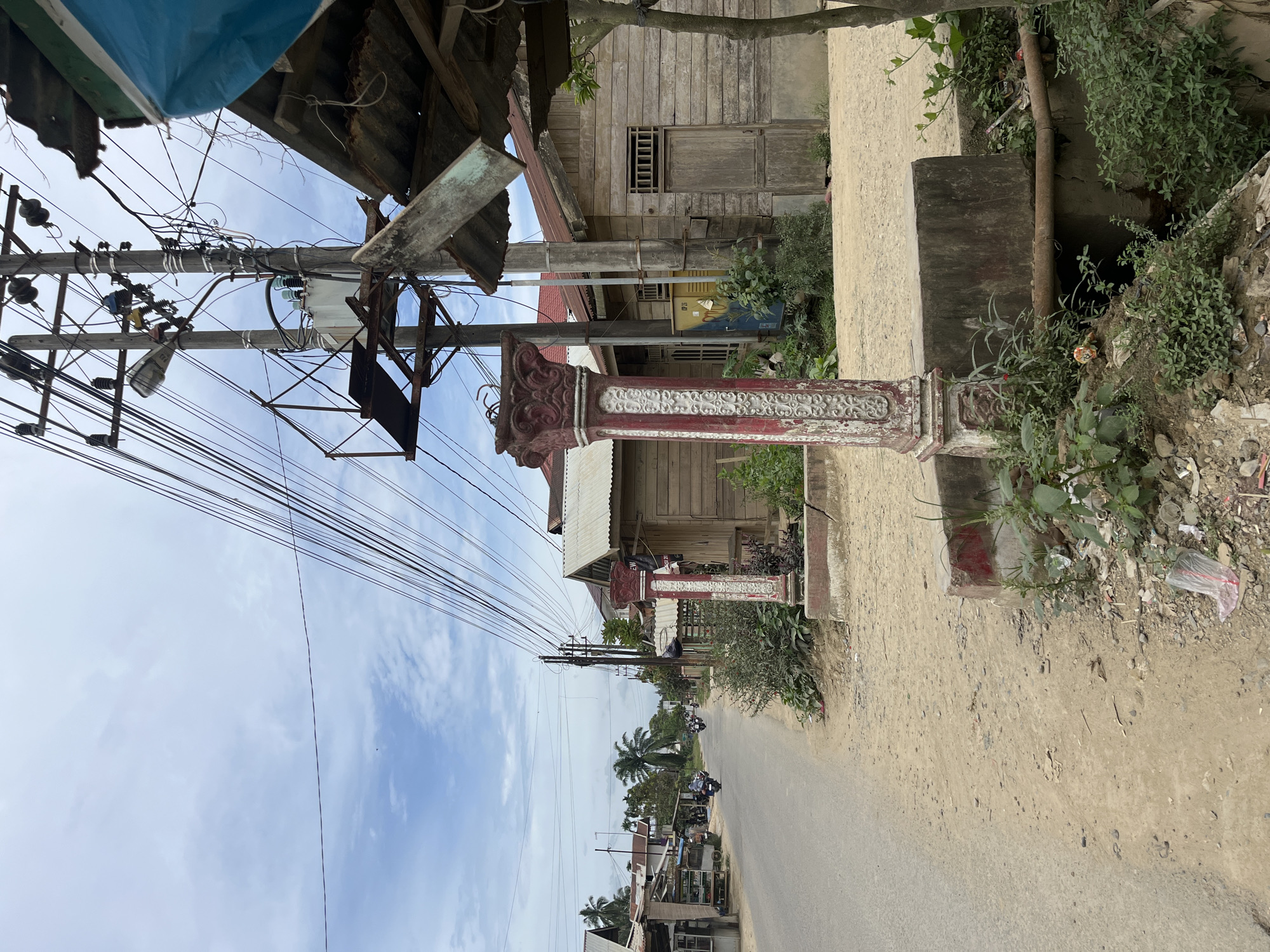A roadside scene in Durian, Indonesia, depicts a paved road running through a village. To the left, a row of small shops with open fronts displays goods, and several motorcycles are parked. Further down the road, motorcycles with riders are visible. On the right side of the road, a wooden house with a corrugated metal roof stands. A decorative red and white pillar is positioned in the foreground, near a concrete structure and an open drain with a metal pipe. Overhead, a dense network of electrical wires and a utility pole with a transformer dominate the sky. Vegetation, including trees and bushes, is present along the roadside and near the structures. The sky is cloudy with patches of blue.
