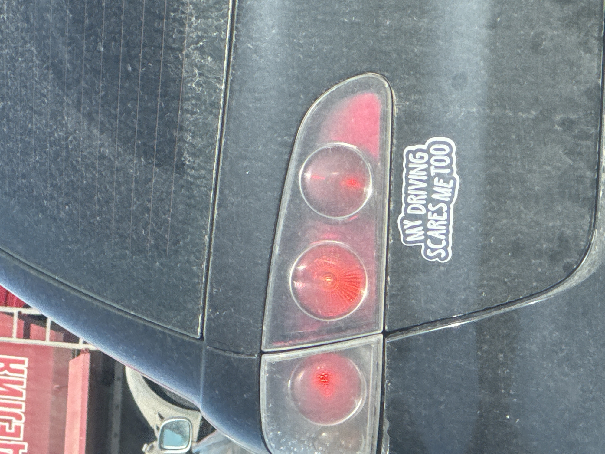 The image shows the rear of a dark grey or black vehicle, likely a car, with a tail light assembly. The tail light is partially obscured by dirt and consists of three circular light elements. A white bumper sticker that reads "MY DRIVING SCARES ME TOO" is affixed to the car's body panel adjacent to the tail light. The vehicle's back window is visible, with vertical defroster lines. Reflected in a side mirror is an unidentified sign in Cyrillic script, suggesting a location where Cyrillic is used. The surroundings include a red or pink painted structure, possibly a building. A city location of Burgas, Bulgaria, is mentioned, but no specific clues confirm this.