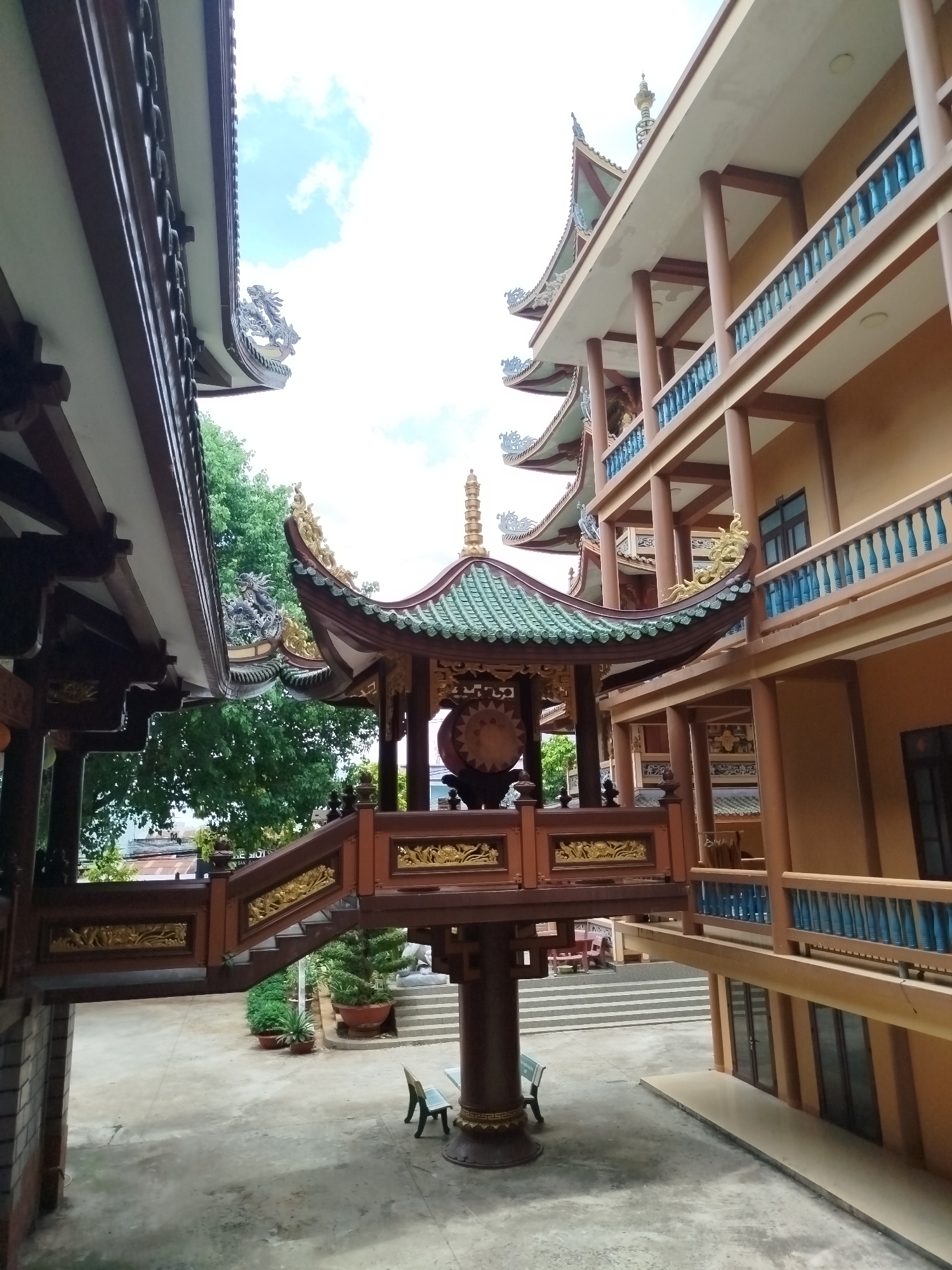 The image captures a Buddhist temple complex located in Pleiku Ward, Vietnam. A two-story building on the right has an ochre-colored facade with blue balustrades. The building has a multi-tiered, pagoda-style roof with green tiles and ornamental golden flourishes.
In the center of the frame stands a small, elevated pavilion. It has a green tiled roof with upturned corners, ornate golden details, and a large drum inside. The pavilion is connected to structures on both sides by short staircases with ornate wooden railings. The left side structure is a darker wood color.
The ground is paved with concrete. There are benches below the central pavilion. In the background, there are stone steps and landscaping with potted plants. The sky is visible above the structures, showing a few clouds.