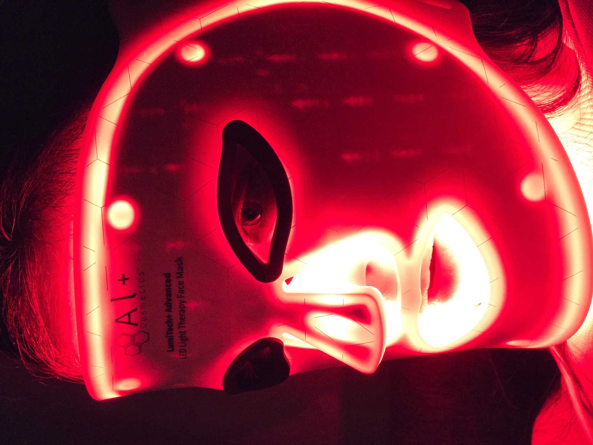 A person wearing an illuminated red LED facial mask is depicted. The mask features cutouts for the eyes and nose, and the mask's brand and model ("AI+ cosmetics LumiTech+ Advanced LED Light Therapy Face Mask") are visible on the side. The person's hair is visible at the sides of the mask. The environment is dark, enhancing the glow of the mask. The location is Unknown, Unknown.