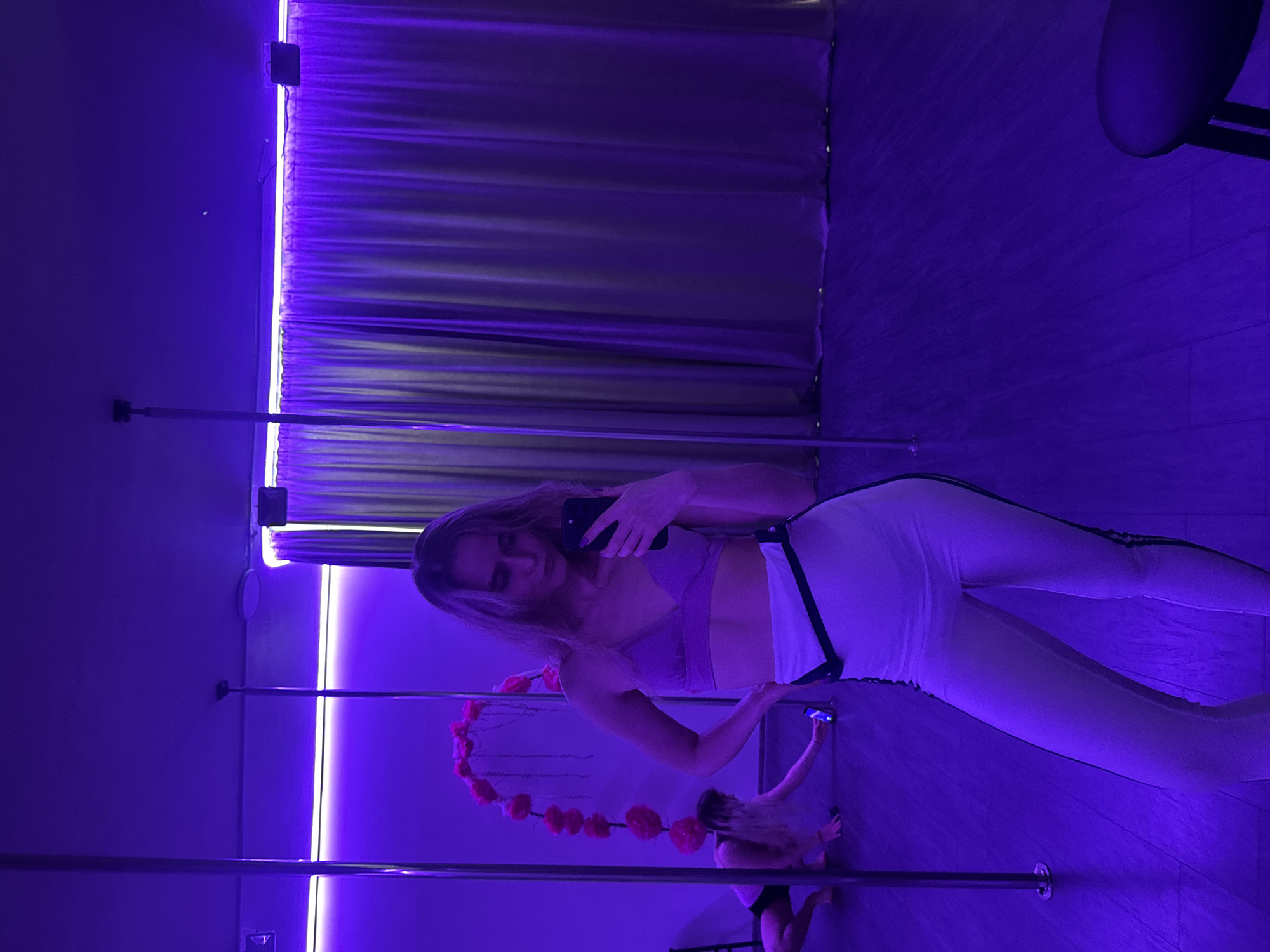 The original image is rotated 90 degrees clockwise, depicting an indoor scene, likely a pole dance or fitness studio, characterized by dramatic purple ambient lighting.
A young woman with long, light-colored hair is the central subject, standing and taking a mirror selfie with a black smartphone. She is dressed in a light-colored sports bra or crop top and matching leggings with dark stripes running down the sides. Her reflection is clearly visible in the large mirror that constitutes much of the background.
In the lower right portion of the scene, another person with light hair is visible, possibly a child or someone further back, also holding a phone with its flashlight illuminated, seemingly looking towards the main subject or the mirror.
The studio features several metallic poles, commonly used for pole dancing. A decorative string of artificial red flowers, possibly arranged in a heart shape, hangs between two of these poles, adding a touch of contrast to the utilitarian equipment and cool lighting.
The dominant visual element is the strong purple and blue light emanating from vertical LED strips along a wall, casting deep shadows and bathing the entire room in this distinct hue. Dark curtains are visible behind the illuminated strips. The floor appears to be a light-colored wood or similar material. A dark chair or stool is partially visible in the upper right corner.
The atmosphere is one of practice or a casual photo session within a dedicated fitness space, highlighted by the striking purple ambient lighting. No discernible text is visible in the image.