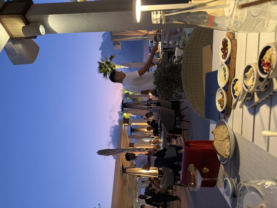 The image depicts an outdoor dining establishment during twilight, likely a high-end restaurant or beach club located in Dubai, United Arab Emirates. The foreground features a white slatted table set with a grey fabric placemat. On the table, a clear cocktail glass with a lime wedge, a red leather handbag with gold hardware, and a white bowl containing sliced rustic bread are visible. A wooden serving tray holds three small white dishes containing olives, olive oil, and a prepared food item. Another white serving tray displays four small white bowls with various dips, one topped with pomegranate seeds. A partially visible clear glass bottle labeled "Evian" and a white, rectangular table lamp are also present.
The midground shows an active dining area with multiple patrons seated at tables featuring wicker chairs. White overhead structures and several large, folded white umbrellas provide partial cover. Artificial lighting emanates from downlights on the white pillars, decorative string lights in the background, and multiple outdoor heating units that cast a warm glow. Two male waiters, dressed in white t-shirts and dark trousers, are performing duties. One waiter, walking from left to right, carries a serving tray with his right arm extended towards a table. Another waiter walks from right to left. A man in a dark suit walks across the far left background. Greenery, including potted shrubs and palm trees, is interspersed throughout the scene.
The background features a twilight sky, transitioning from a deep blue at the top to hues of purple and orange-pink near the horizon, indicative of sunset. Scattered clouds are visible against the colorful sky. A distant, faint horizon line is discernible, suggesting a body of water or landmass beyond the dining area.