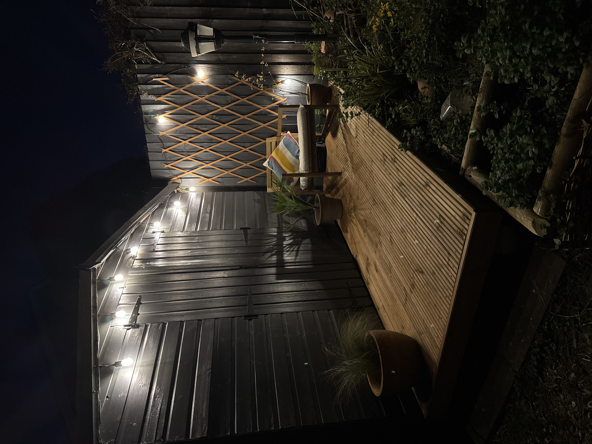 The image depicts an outdoor space in South Kesteven, United Kingdom at night. The area is illuminated by string lights hanging on a black shed and a light fixture on a fence. A wooden deck extends from the shed, furnished with a wooden chair adorned with pillows. Two potted plants flank the deck and chair. The scene is enclosed by a fence and vegetation.