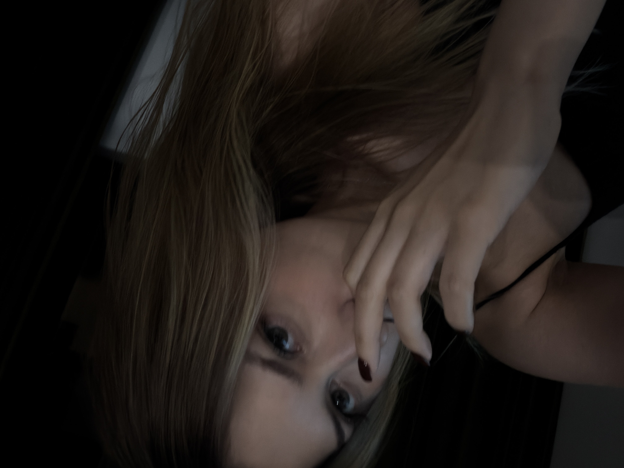 The image is a close-up, low-light selfie of a young woman with long, blonde hair. She is wearing a black garment, possibly a tank top, and her left hand is raised to her face, with her fingers touching her nose. Her eyes are open and she is looking towards the camera. The background is dark and indistinct. There is no indication of a specific location within the city of Đà Nẵng, Vietnam.