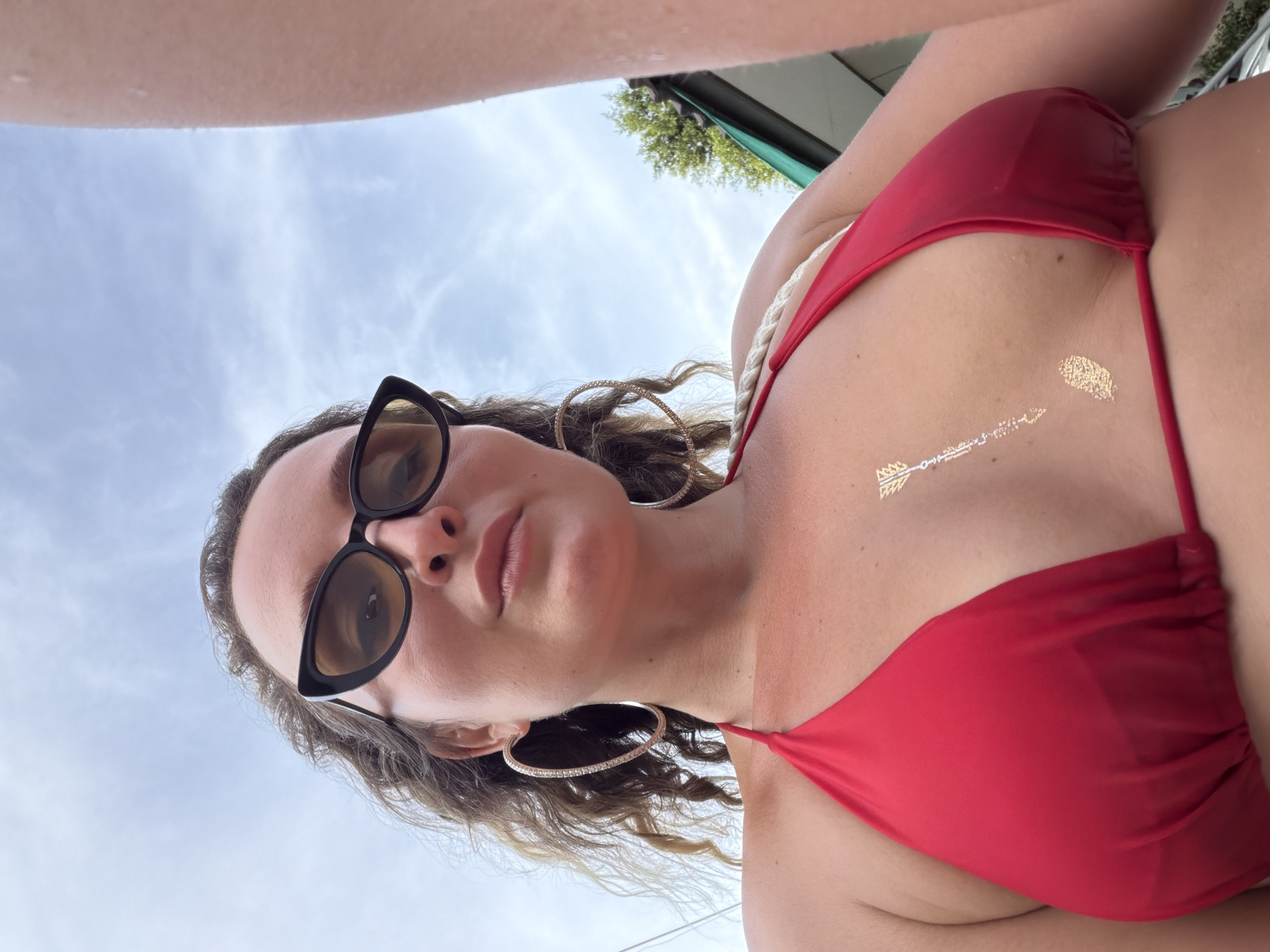 In a low-angle, close-up shot taken outdoors, a woman wearing a red bikini and dark, cat-eye sunglasses is seen from the chest up against a cloudy sky. Her curly, brown hair frames her face, and large, hoop earrings adorn her ears. A metallic, temporary tattoo of an arrow and a feather is visible on her chest. The background shows glimpses of green foliage and what appears to be a structure, suggesting an outdoor setting. This image was taken in the Ko Pha-ngan District, Thailand.