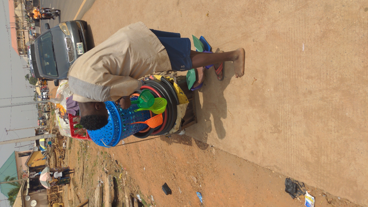 This outdoor image, taken in bright daylight, depicts a street scene in what appears to be an urban or semi-urban area, consistent with Ado, Nigeria.
In the foreground, a person, likely a young male, is bent over a makeshift display or cart laden with various items for sale. He wears a light-colored long-sleeved tunic over dark blue shorts. His feet are bare, with one foot showing a red-brown sandal and another pair of green flip-flops nearby. The cart holds a prominent blue plastic laundry basket or basin, containing and surrounded by colorful plastic kitchenware such as green, yellow, and orange sieves, strainers, and scoops. Coiled black hoses or tubing are also part of the merchandise. The person's posture suggests he is attending to his wares, possibly arranging them or looking into the basket.
Behind the vendor, a grey sedan is visible on a paved road, displaying a Nigerian license plate that reads "KLJ-956R." Further down the road, a person on a motorcycle is seen, alongside other vehicles and pedestrians, indicating a busy thoroughfare. The ground immediately around the vendor is a mix of light brown dirt and a lighter paved surface.
The background shows a bustling streetscape with various buildings, some featuring corrugated roofs, and what appears to be market stalls or small shops. Utility poles and power lines are visible against a clear, bright sky, suggesting midday or early afternoon. The overall setting is that of a vibrant commercial street. Scattered litter, including a black plastic bag and wrappers, is visible on the ground.