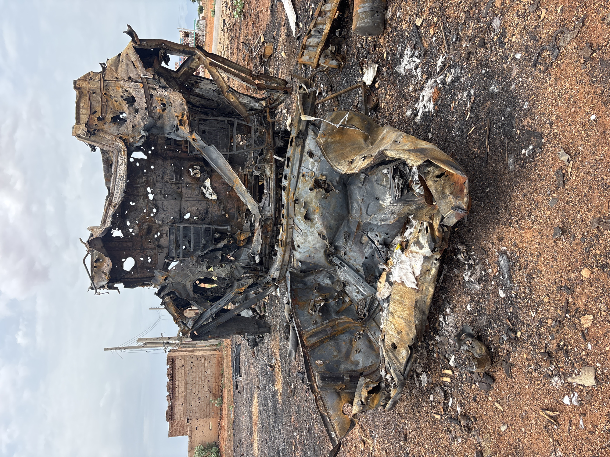 The image depicts a scene of destruction in Um Laba, Sudan. In the foreground and mid-ground are the heavily charred and mangled remains of what appears to be a vehicle, likely an SUV or van, completely destroyed by fire. The metal is twisted, melted, and blackened, with numerous holes and gaps visible, suggesting the intensity of the blaze and possible impacts. Some white, fibrous material, possibly insulation or debris, is scattered around the wreckage. The ground is covered in reddish-brown soil, ash, and scattered debris from the destruction.
In the background, a damaged brick building with visible holes, likely from gunfire or shrapnel, stands to the left. A utility pole with electrical wires is also visible. Further in the distance, other damaged structures and the landscape of Um Laba can be seen under a cloudy sky. The overall impression is one of recent conflict or disaster, with the destroyed vehicle being the central subject of the image. No people are visible in the frame.