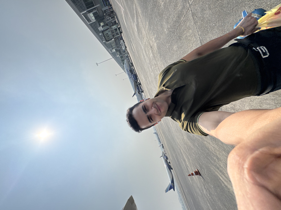 A man smiles in a selfie on an airport tarmac. He wears a dark green polo shirt, black shorts, and a gold bracelet. In his right hand, he holds a shopping bag. Behind him are several parked airplanes, including one with "IndiGo" visible. The airport building is in the background. The sun shines brightly in the sky.