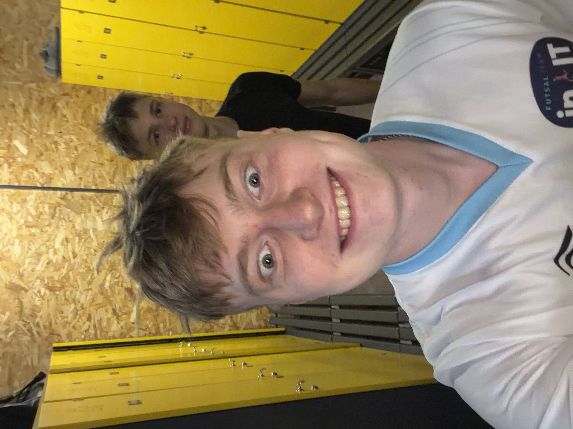 A selfie taken in a locker room shows two young men. The man in the foreground, who is taking the photo, is smiling widely with his eyes wide open. He is wearing a white t-shirt with a blue trim around the collar. Behind him, another young man in a black t-shirt is partially visible, looking at the camera. The background features rows of yellow lockers and wooden-paneled walls. The scene appears to be inside a sports facility.