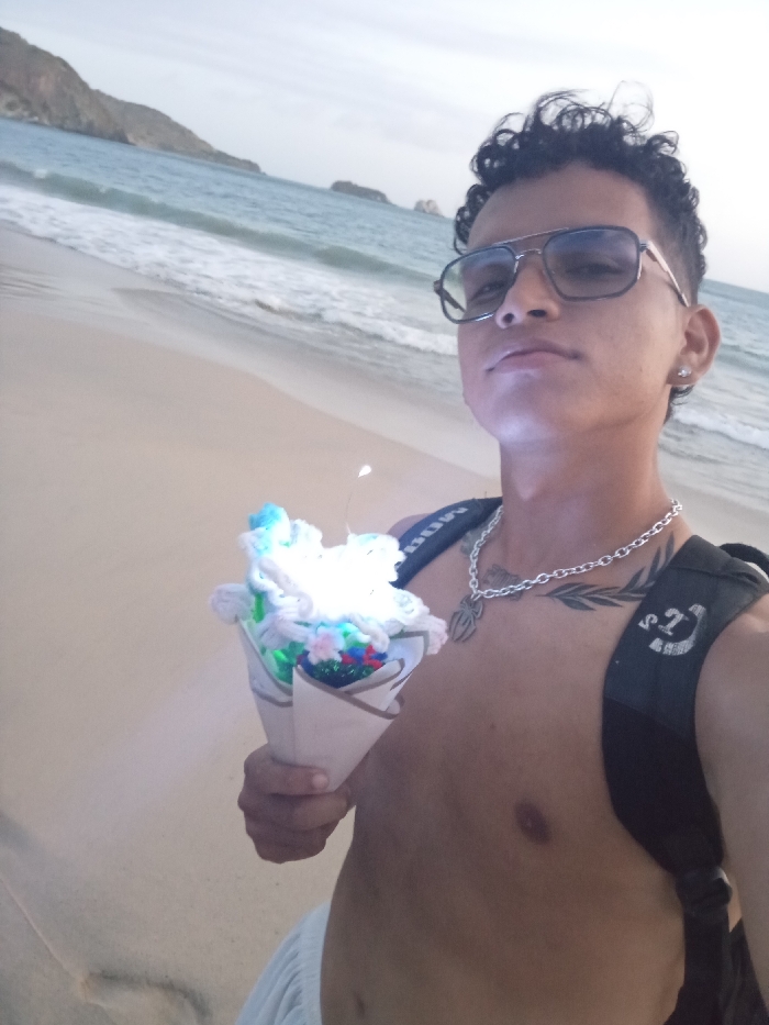 A shirtless man with a backpack and sunglasses stands on a beach, holding a cone containing fluffy, glowing materials and flowers. He wears a silver chain with a spider pendant. A tattoo is visible on his chest. The beach stretches to the ocean, with distant rocky formations and a cloudy sky. Waves are visible near the shoreline.