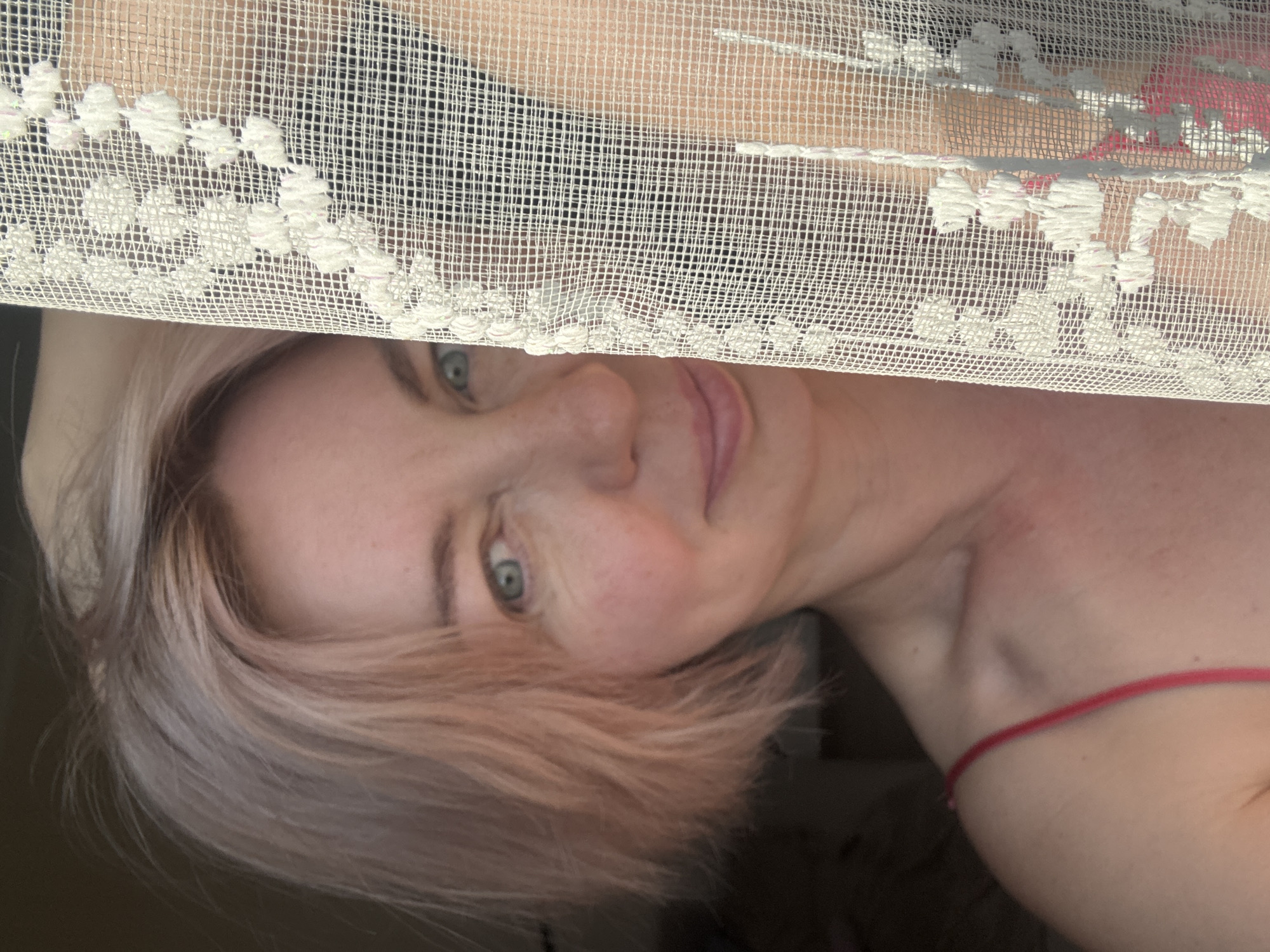 The image shows a close-up, slightly tilted portrait of a woman looking directly at the camera. Her face, including her light blue-green eyes, nose, and mouth, is partially visible, peeking from behind a sheer, light-colored fabric that covers the upper portion of the frame. This fabric has a mesh-like, woven grid pattern and features white embroidered details along its edge, resembling lace or tulle.
The woman has short, light-colored hair, possibly blonde or light brown, framing her face. Her expression is neutral to slightly smiling. A part of her bare shoulder is visible on the right side of the image, with a thin red strap, possibly from a garment or accessory. The background behind the sheer fabric is indistinct, appearing as soft tones, suggesting an indoor setting with diffused lighting. No specific time of day or weather can be determined from the image, though the lighting suggests daytime. There is no visible text in the image. The image is set in Sochi, Russia, according to the provided context.