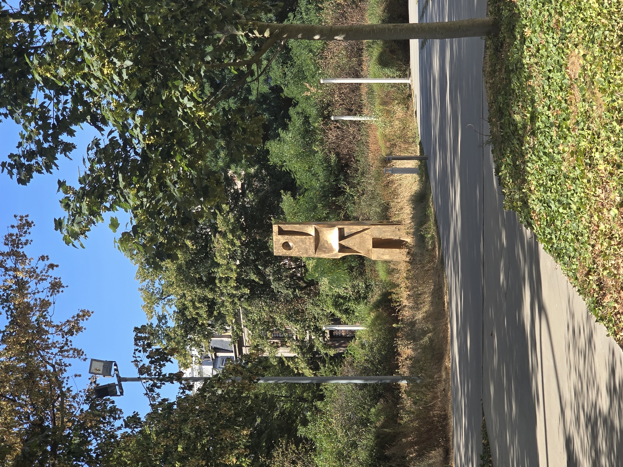 The image depicts an outdoor scene in Antwerp, Belgium, featuring a modern sculpture. The sculpture is tall and abstract, made of a golden material, with cut-out shapes and geometric forms. It is positioned in a landscaped area with dry grasses and bushes. In the background, a building with visible windows and a pitched roof can be seen amidst dense greenery. To the left of the sculpture, a tall pole with two industrial-style lights is visible, also surrounded by trees. The foreground shows a paved pathway or road with dappled shadows cast by overhead trees. The sky is clear and blue. There are no people visible in the image. The overall environment suggests a park or public urban space.