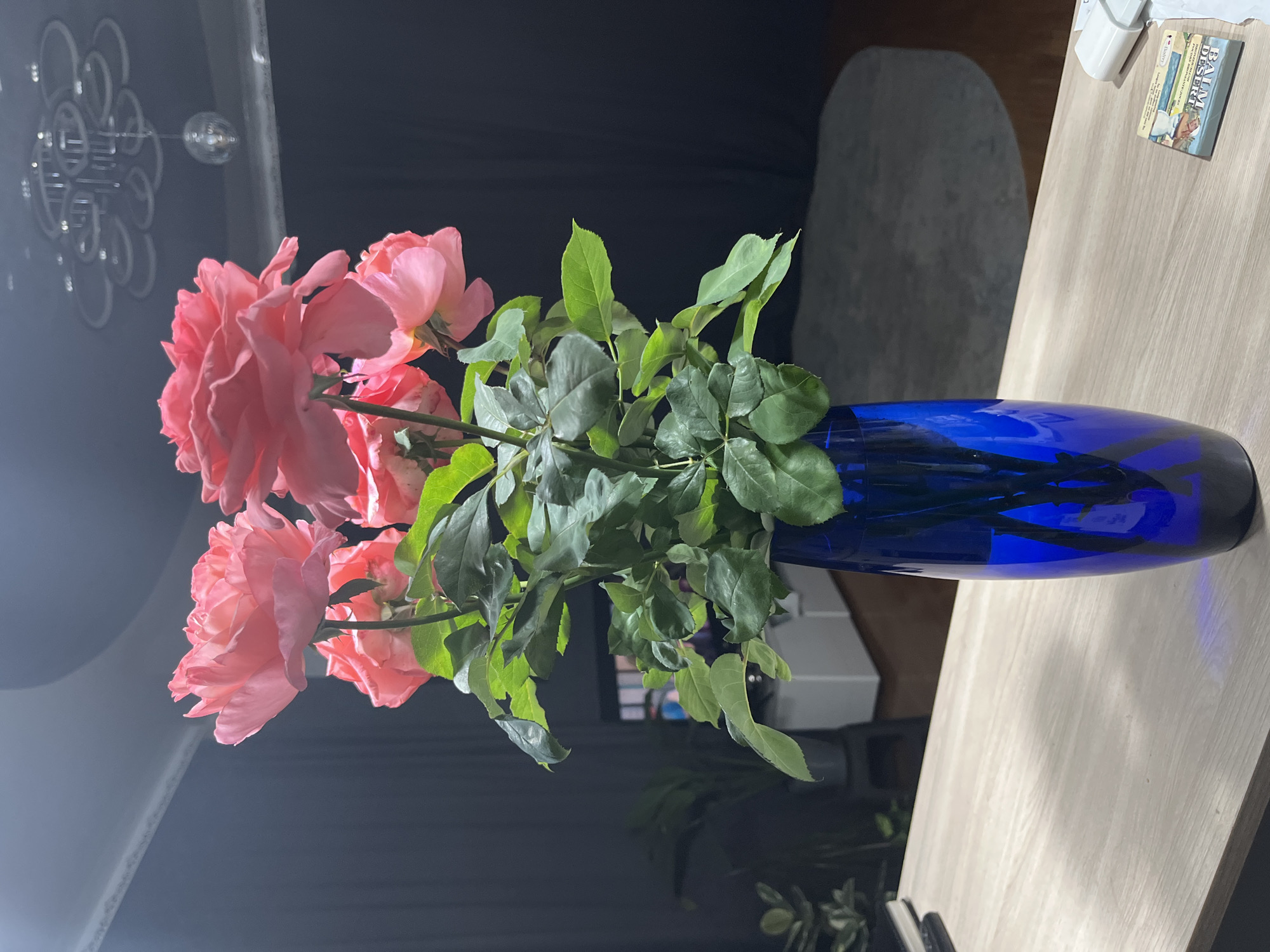 A bouquet of bright pink roses in a cobalt blue glass vase sits on a light wood-grain table. The roses have large, fully bloomed heads and are surrounded by dark green leaves. Behind the roses, a dark grey curtain is drawn. In the background, a white cabinet is visible, along with a television screen. Above the curtain, a decorative ceiling fixture with multiple rings is illuminated, and a small disco ball hangs from its center. The lighting is dim, creating shadows on the wall and floor. The scene appears to be indoors. The image does not provide any information regarding a specific location in Saratov, Russia.