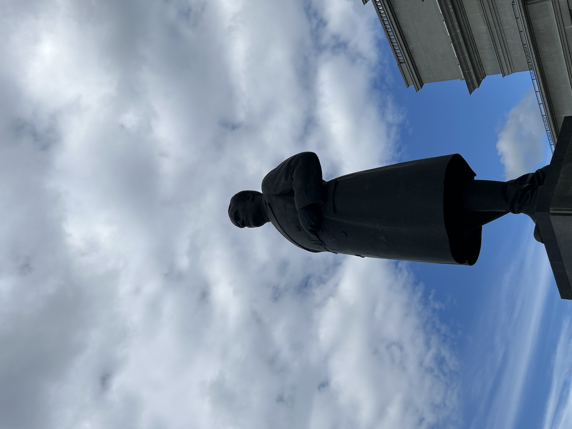 A low-angle shot captures a dark statue of a man in a long coat standing against a cloudy blue sky. The statue is positioned on a dark pedestal, and its left side is angled towards the viewer. To the right of the statue, a portion of a large, grey, multi-story building with visible architectural details and a railing is visible. The sky is filled with a mix of white and grey clouds, with patches of blue sky peeking through. The image appears to be taken outdoors in a public space. This is a statue of Ivan Ivanovich Nesterov, located in Novokuybyshevsk, Russia.