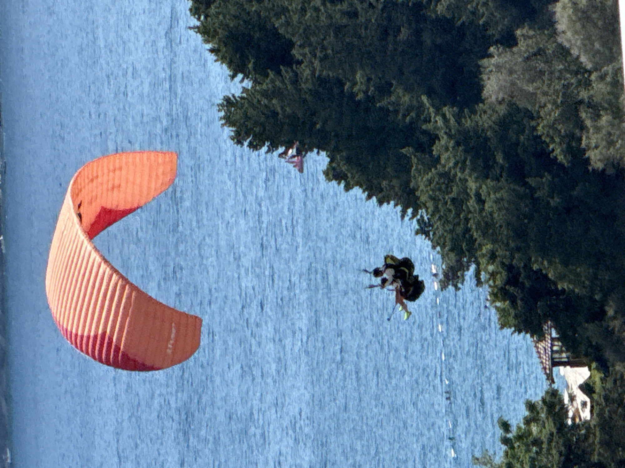 An aerial shot captures a paraglider soaring above the blue waters of the Adriatic Sea, near Boreti, Montenegro. The paraglider, a vibrant orange and red canopy, is piloted by two individuals. They are suspended in a harness below the wing, appearing to be a tandem paragliding experience. The paraglider is positioned in the upper left quadrant of the image, with the vast expanse of the sea filling most of the frame.
In the lower right section of the image, a dense cluster of dark green trees forms a stark contrast to the bright blue water. A small, red object, possibly another paraglider or a drone, is visible in the distance over the water, near the treeline. Below the trees, a partially obscured tiled roof of a building can be seen. In the middle ground, a line of small, white buoys stretches across the water, suggesting a marked swimming or boating area. The overall scene depicts a recreational activity taking place in a coastal environment.