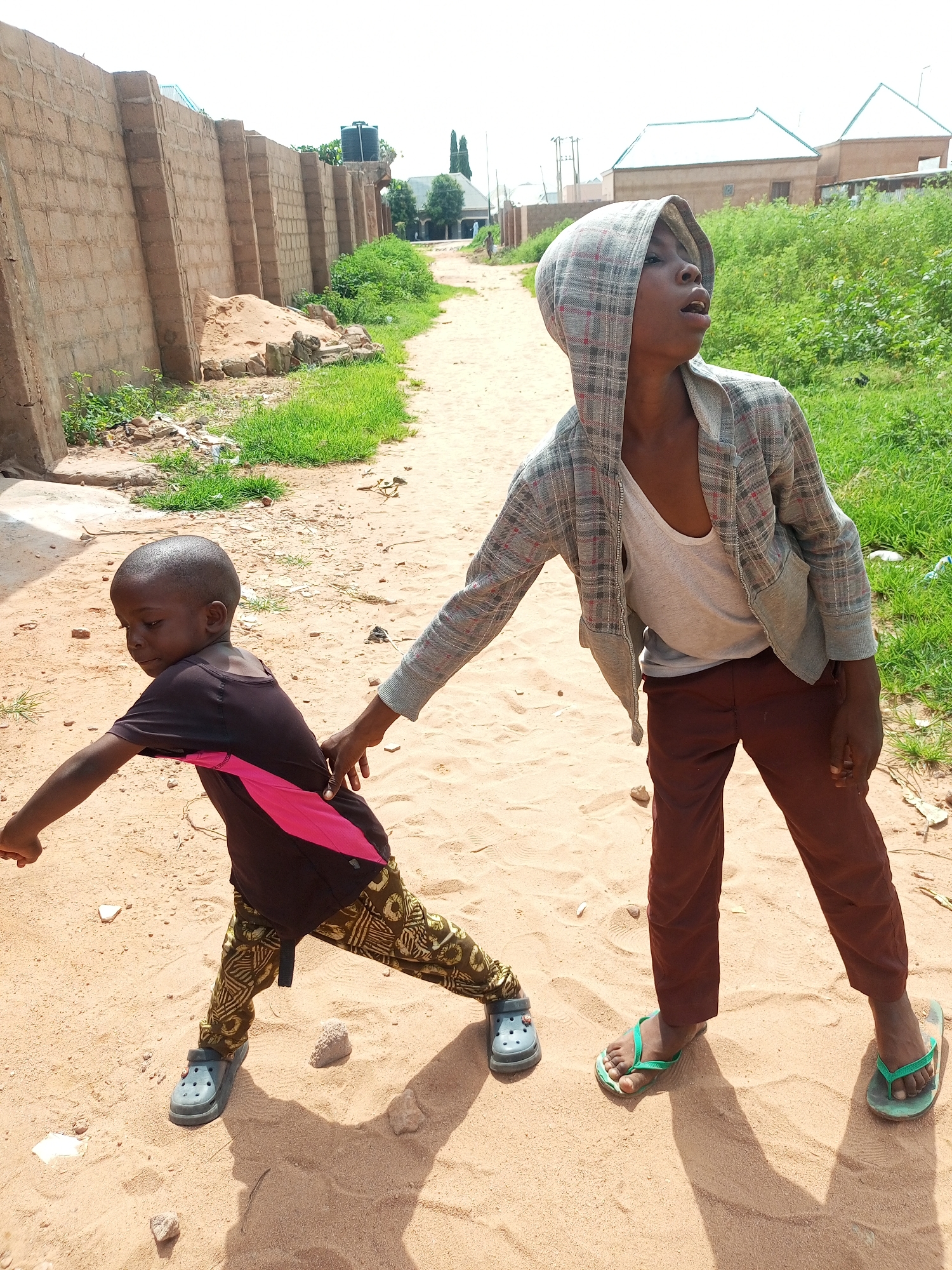 In Tempure, Nigeria, two young boys are captured in a dynamic pose on a sandy path. The boy in the foreground, on the left, is wearing a dark t-shirt with a vibrant pink accent and patterned trousers, along with gray clogs. He is leaning forward, his right arm extended, as if reaching or running. Behind him, the second boy, taller and wearing a hooded checkered jacket over a white tank top and brown pants, with green flip-flops, has his hand on the first boy's back, guiding or restraining him. The path they are on is sandy and appears to be a common thoroughfare, bordered by a brick wall on the left and green vegetation on the right. In the background, simple houses with distinct rooftops are visible, suggesting a semi-urban or rural setting within Tempure. The lighting indicates it is daytime, with shadows cast on the sand.