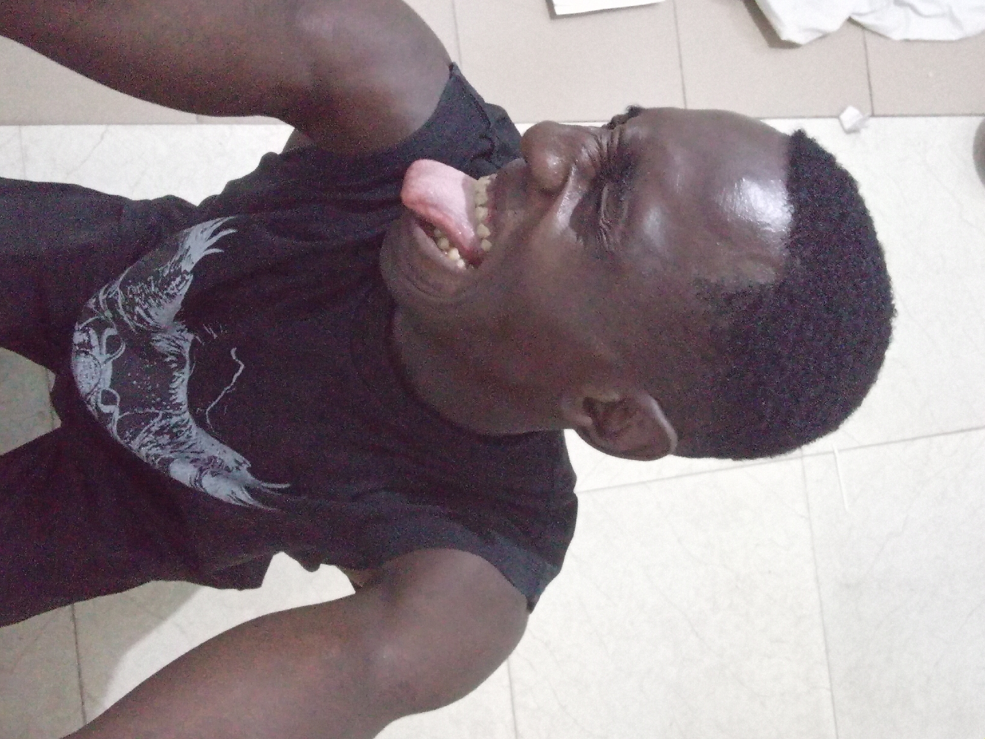 A top-down perspective captures a dark-skinned individual wearing a dark t-shirt with a white graphic design, possibly a bird, on the front. The person is sticking their tongue out with their eyes squinted or closed. The background reveals a tiled floor, possibly indoors. There is no context to confirm if this is in Akwete, Nigeria.