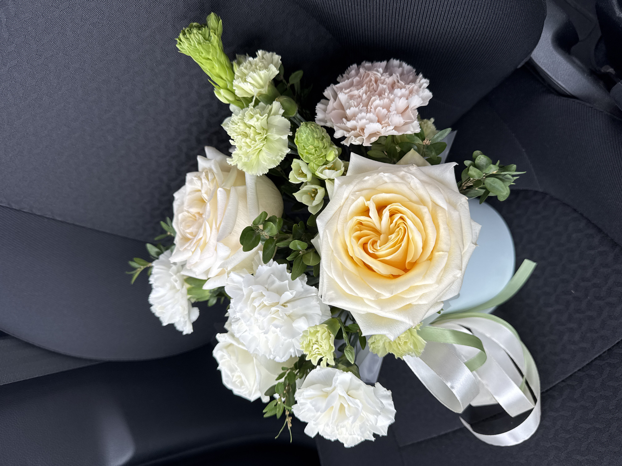 A vibrant bouquet of fresh flowers rests on a dark gray or black car seat, captured in Izhevsk, Russia. The prominent subjects are several roses, primarily creamy white, with one large rose featuring a striking yellow-orange center. These are complemented by ruffled carnations in white, light green, and a soft blush pink hue. The arrangement also includes spiky green filler flowers, possibly Ornithogalum, and small green leaves, adding texture and depth.
The flowers are artfully contained within a small, light blue or mint green circular base, from which delicate white and light green ribbons extend. The background is distinctly the textured fabric of car upholstery, suggesting the bouquet is in transit or has just been placed there. Hints of the car's interior plastic trim are also visible. There are no people or discernible text in the image. The lighting appears natural and bright, enhancing the fresh appearance of the flowers. The scene conveys a sense of anticipation or completion, as the bouquet awaits its final destination or recipient.