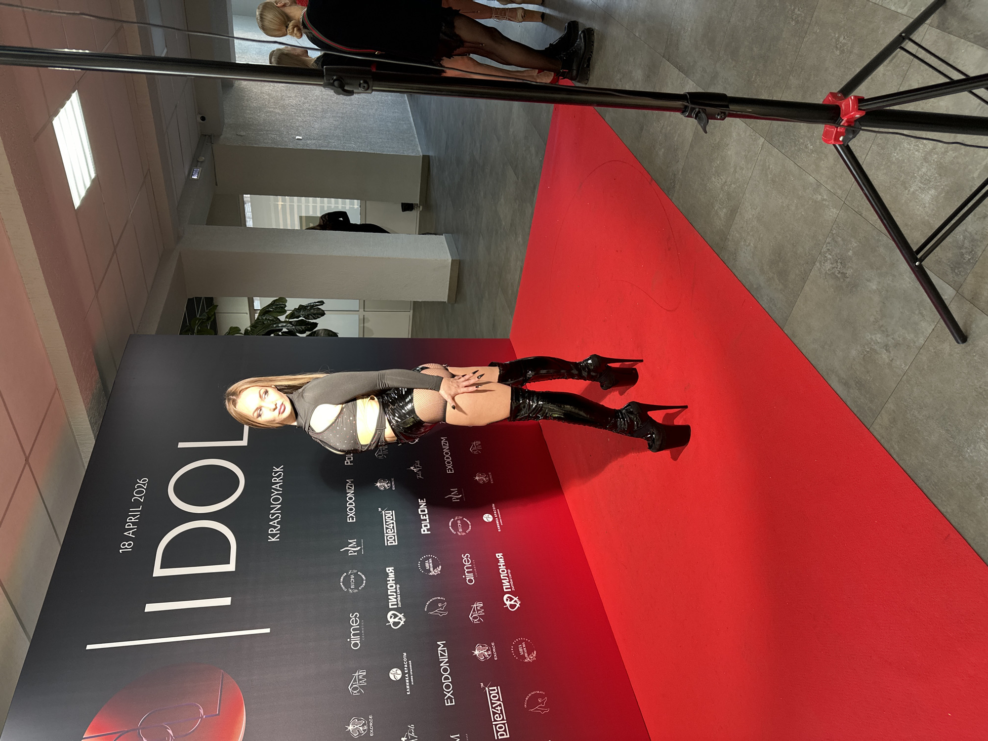 A woman is posing on a red carpet in front of a backdrop promoting an "IDOL" event scheduled for April 18, 2026, in Krasnoyarsk. The backdrop also features various logos of sponsors including "EXODONIZM", "ISTOMA", "aimes", "PM", "pole4you" and "POLEONE". The woman is wearing a cutout top, shiny black shorts and black thigh-high stiletto boots. There is a black light stand in the shot, and other people can be seen in the background. The floor is made up of grey tiles.