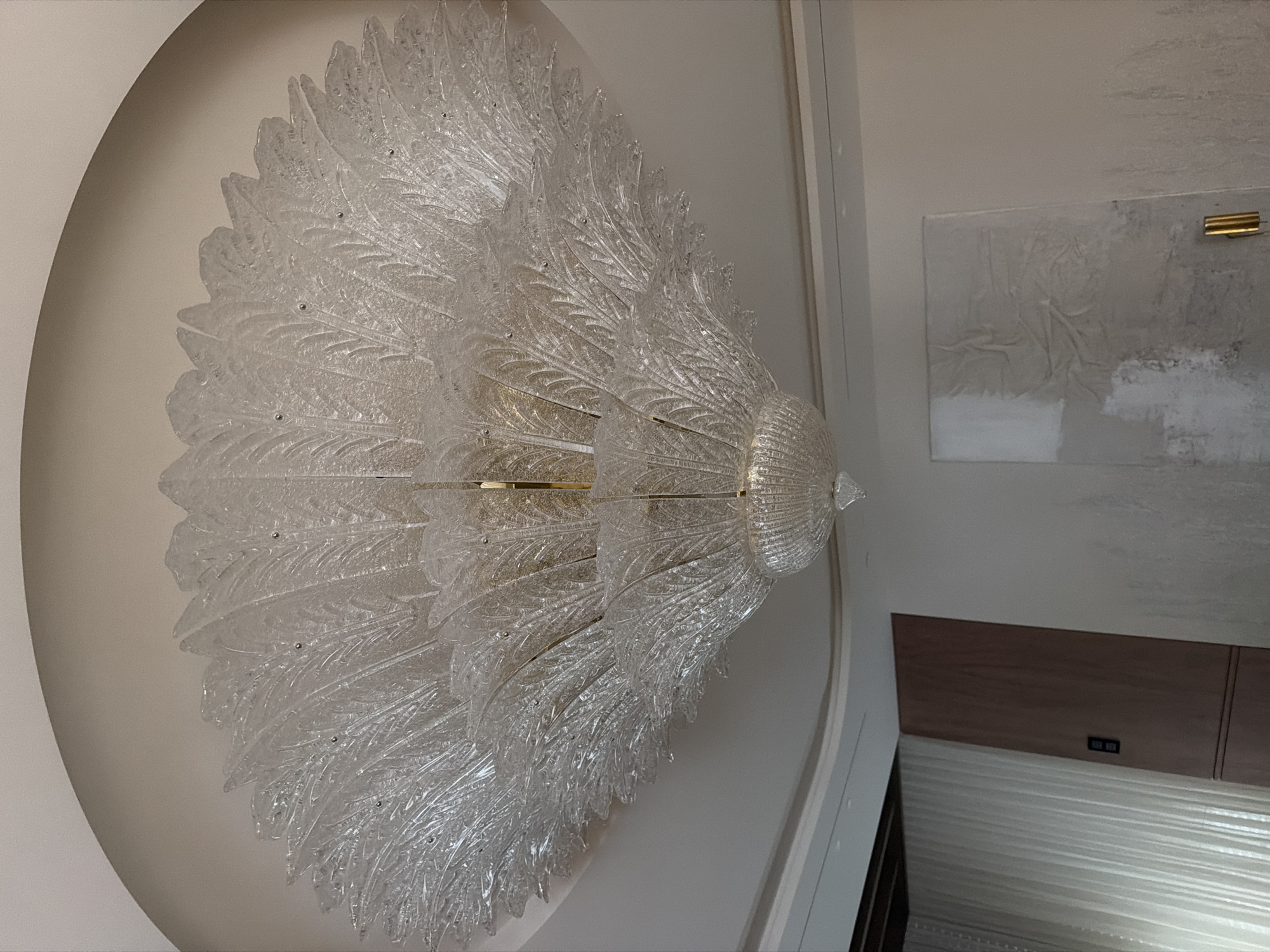 A large, ornate chandelier made of clear, textured glass that resembles feathers hangs from a white ceiling. The chandelier has multiple tiers and appears to be quite elaborate. Below the chandelier, on the right side of the image, a portion of a wall is visible with a textured artwork hanging on it. A vertical gold accent is also present on the wall below the artwork. On the left side of the image, a tall, dark wood cabinet or door is seen next to a wall covered with sheer white curtains. The overall scene depicts an interior space, likely a room in a luxury establishment, though its specific location in Moscow, Russia cannot be determined from the image alone.