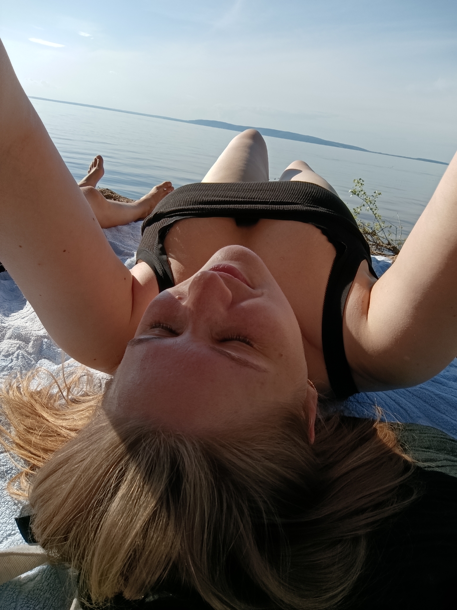 A woman is lying on her back on a towel by a body of water. She is wearing a black tank top and her eyes are closed. Her arms are raised above her head. Her hair is blonde and spread out around her head. The water is calm and there is a light blue sky with some clouds. The scene appears to be outdoors in a natural setting.