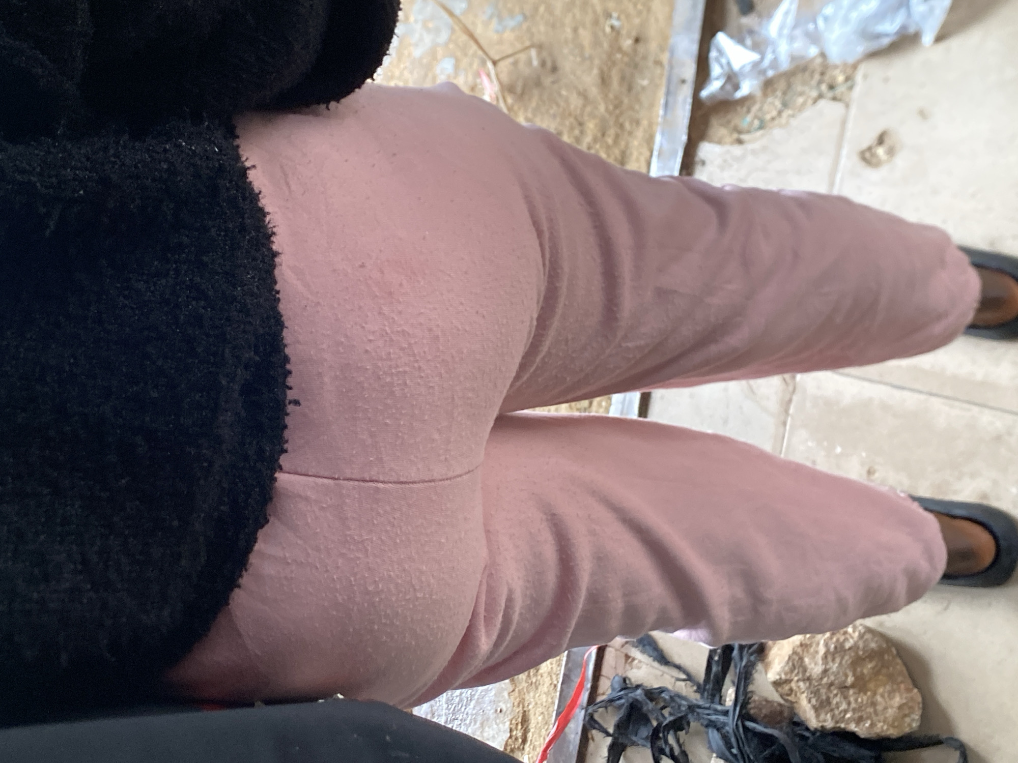 A close-up overhead shot shows a person wearing pink sweatpants and black shoes. The person is also wearing a black fuzzy garment. The background consists of a tiled surface with debris and a plastic bag. The location is not discernible from the image and no details suggest the city of Bukuru, Nigeria.