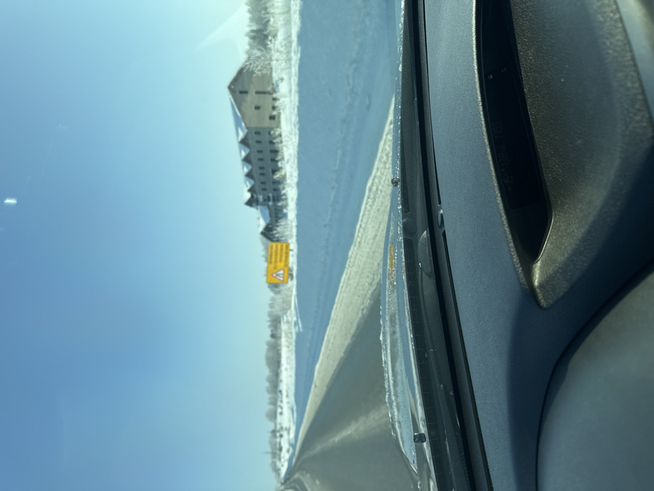 A view from inside a vehicle reveals a snow-covered landscape. A road runs alongside a snow-laden field, bordering a building complex. A yellow warning sign stands prominently along the roadside. A clear, blue sky dominates the upper portion of the frame. The right side showcases the interior of the car, including part of the dashboard and windshield.