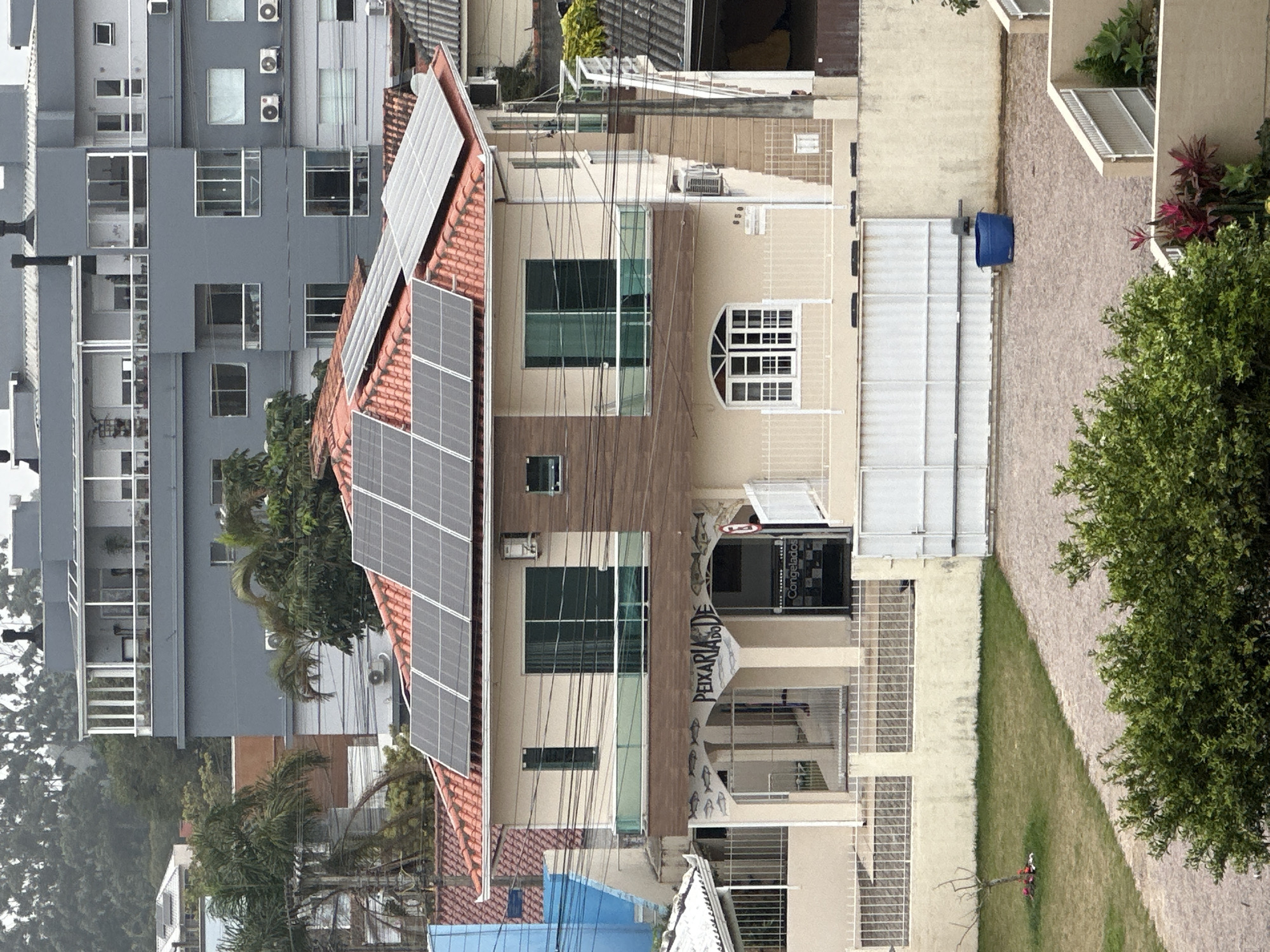 This image depicts a two-story building with a tan facade, featuring a red-tiled roof topped with solar panels. The building's lower level has a storefront with large windows and the text "PEIXARIA DE" visible, indicating it is a fish market. A white fence encloses the property, and a large white gate is present. To the right of the building, there is a gravel driveway with a blue plastic barrel. In the foreground, a green lawn leads to a gravel path, bordered by lush green bushes. In the background, several other buildings are visible, including a larger, grey, multi-story residential building with balconies and air conditioning units. The scene is situated in the city of Florianópolis, Brazil, likely in a residential or mixed-use area. The overall atmosphere is overcast, suggesting a cloudy day. No people are clearly visible in the frame, and no specific actions or interactions are occurring.