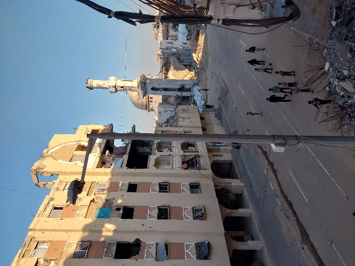 The image depicts a street scene in Khan Yunis, Palestinian Territory, characterized by significant structural damage. In the foreground and left, a multi-story, cream-colored residential building exhibits severe destruction, including large holes, exposed interiors, shattered windows, and a partially collapsed upper section featuring a distinctive arched façade element. Its lower arches are intact, with a vehicle partially visible underneath.
To the right of the street, a tall, grey streetlamp pole with wires extends upwards. In the mid-ground, a group of approximately ten individuals, predominantly males and boys, are gathered on the asphalt road; several appear to be engaged in a game of soccer with a dark ball. Debris and scattered rubble are visible along the road's edges and in the immediate right foreground, including twisted rebar. A paved sidewalk runs along the left side of the street, and a bicycle is parked further down on a sidewalk to the right.
In the background, a mosque with a prominent white minaret and a light-colored dome stands amidst further structural damage and rubble, indicating widespread destruction. The sky is clear and blue, with sunlight illuminating parts of the scene and casting shadows, particularly on the left side of the damaged residential building.