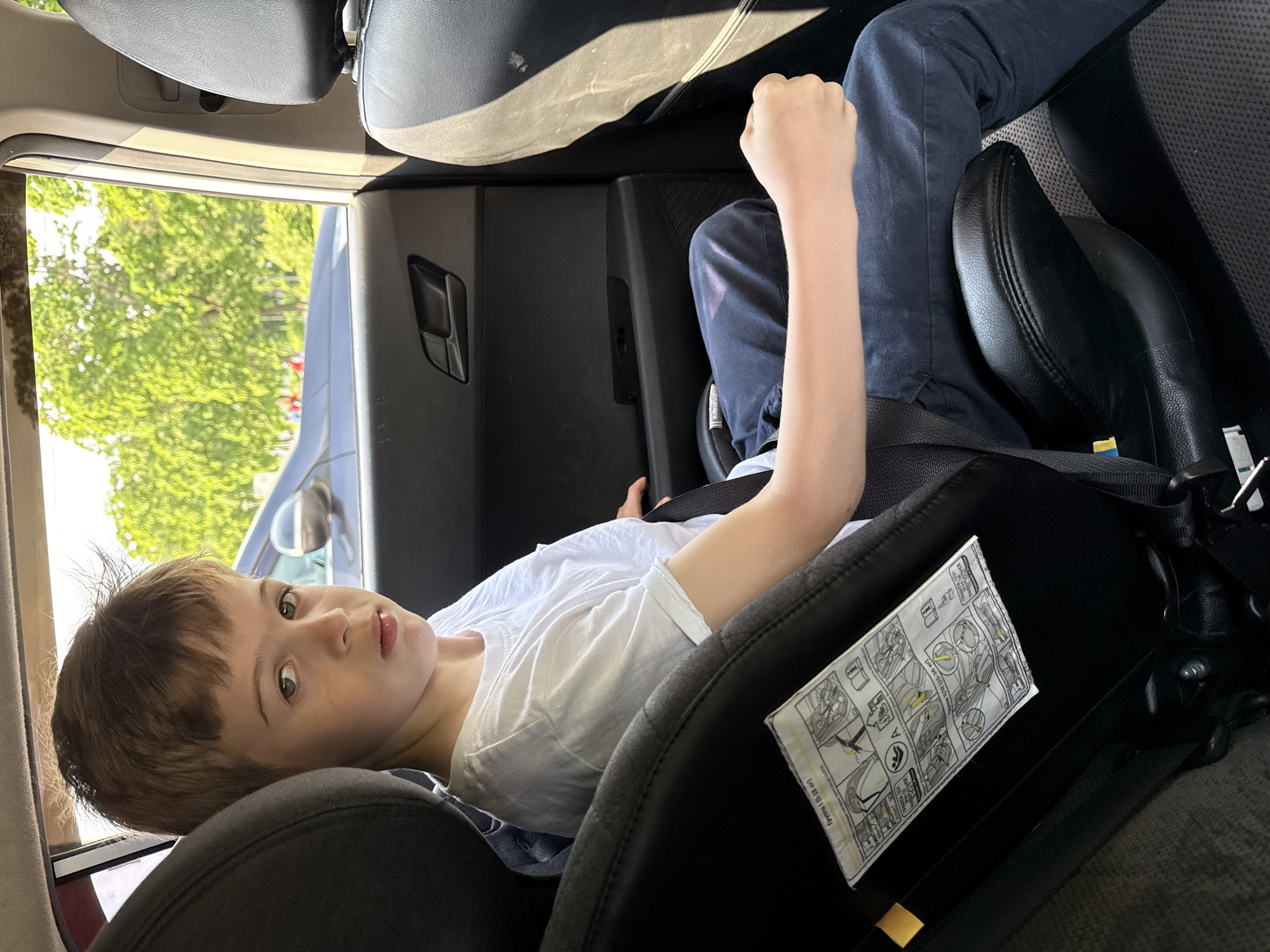 A young boy with brown hair lies in a black car seat inside a vehicle. He wears a white t-shirt and blue pants and has his fist clenched. A white instruction label is attached to the car seat. Outside the vehicle, green trees and a glimpse of another car are visible through the open window. The location of Khimki, Russia, cannot be determined from the visible elements.