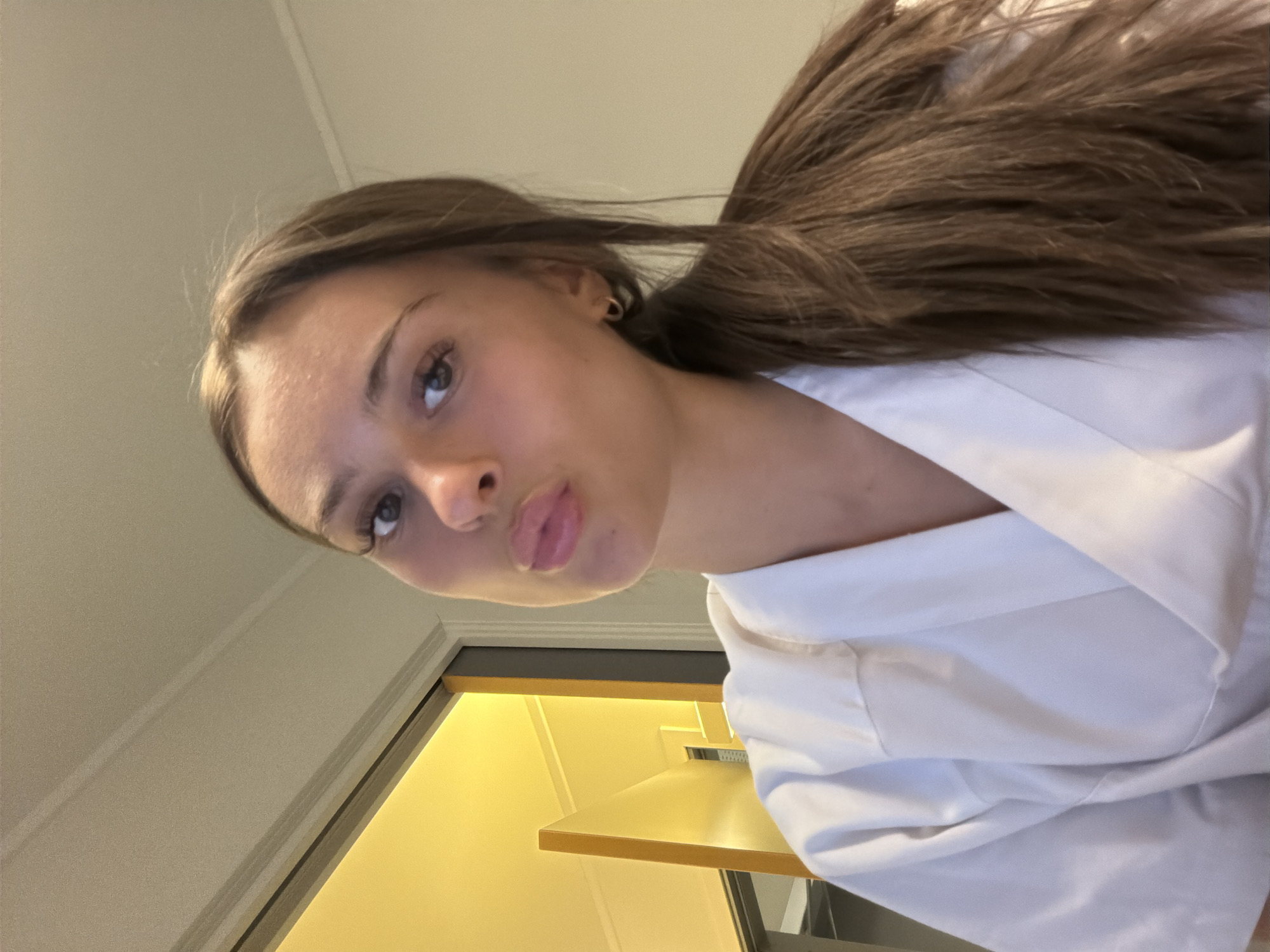 A young woman with long brown hair tied in a ponytail, wearing a white medical scrub top, looks at the camera with her lips puckered in a kiss. She has brown eyes, defined eyebrows, and small hoop earrings. Behind her, a doorway reveals a bright yellow wall and a staircase. The setting appears to be indoors, likely a home or a medical facility, in the city of Harstad, Norway.