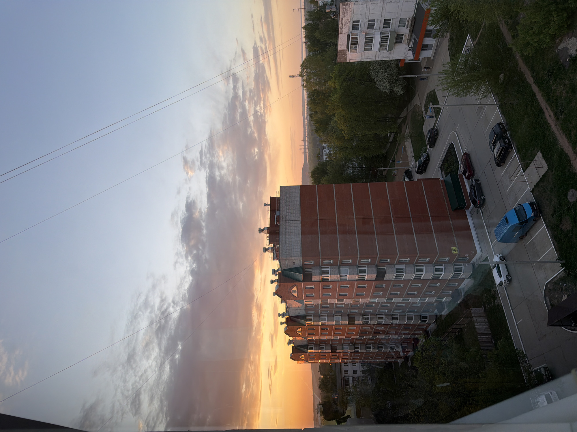 The image depicts a high-angle view of a cityscape in Chaikovsky, Russia, at what appears to be sunrise or sunset. The sky is filled with a gradient of colors, transitioning from a pale blue to a vibrant orange near the horizon, partially obscured by clouds. A tall, brick-faced apartment building dominates the center of the frame, with its multi-storied structure reflecting the warm light of the setting sun. To the right of the apartment building is a parking lot with several parked cars, including black, red, white, and a blue van. Trees and other greenery surround the parking area. Further back, there is another building with a white facade. Overhead power lines are visible against the sky. The photograph appears to be taken from inside a building, looking out through a window.