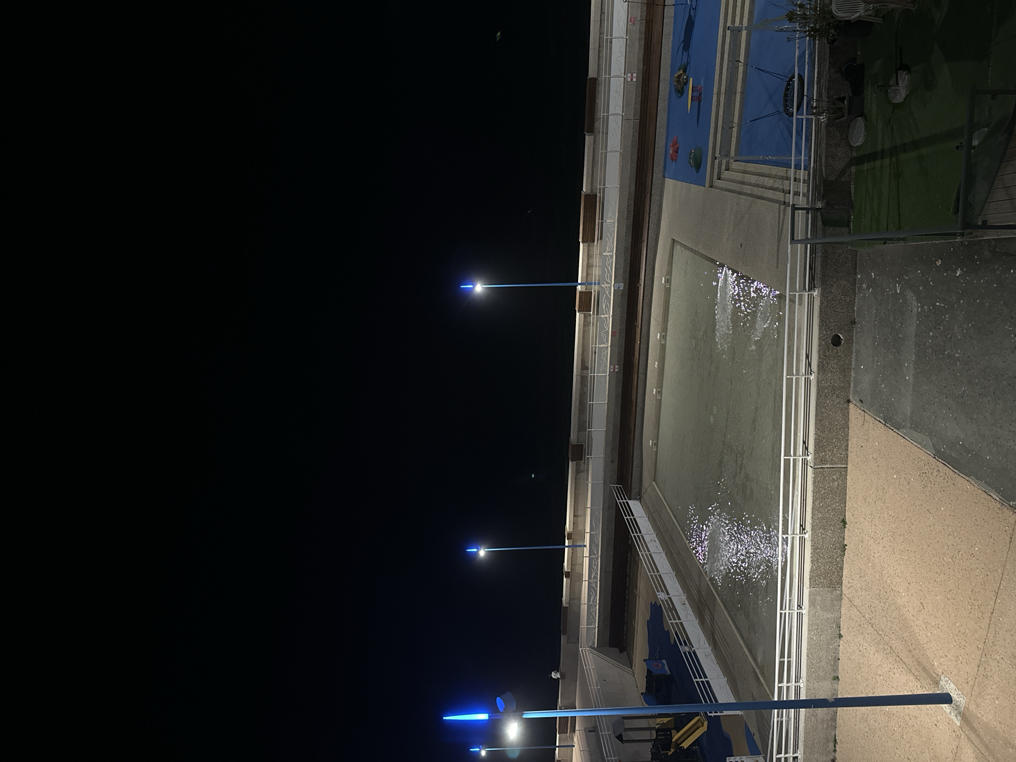 The image shows a night scene of an outdoor swimming pool complex and playground area adjacent to the sea in Saint-Valery-en-Caux, France. The sky is dark, indicating nighttime. Several blue lampposts illuminate the area. The swimming pool is partially filled with water, and the surface reflects the artificial light, creating shimmering patterns. A white railing encloses the pool area. Adjacent to the pool is a blue-surfaced playground with various play structures, including swings and what appears to be a roundabout. Further in the background, a paved walkway runs along the sea wall, lined with wooden benches. The sea is visible as a dark expanse beyond the walkway. There is one person visible, standing near the walkway, and a small white object that could be a bird in flight over the sea. The foreground shows a textured concrete or stone pathway. The overall scene depicts a public recreational area at night.
