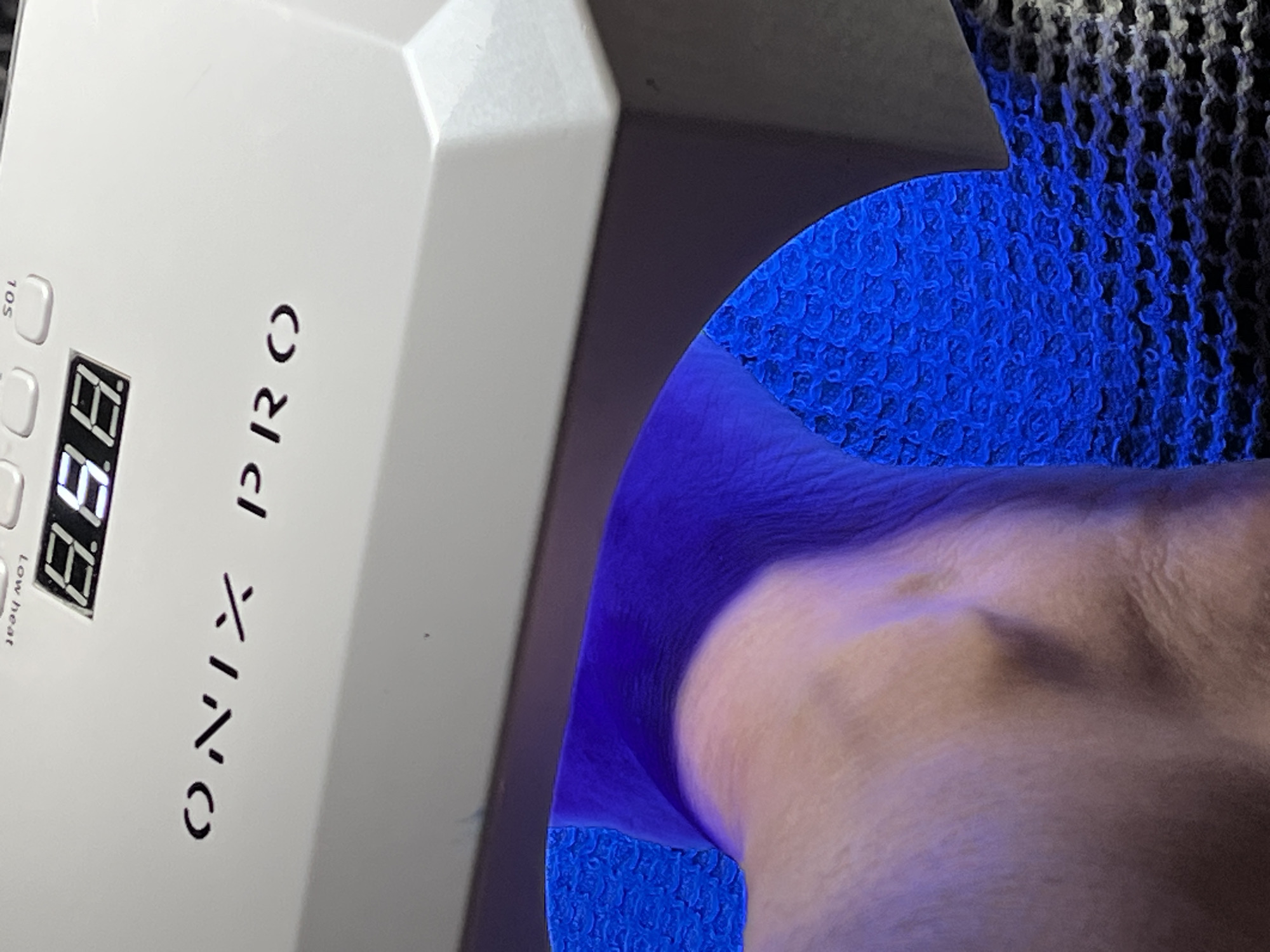 The image shows a close-up of a white UV nail lamp with the brand name "ONIX PRO" printed on it. A digital display on the lamp shows "8.8.". The lamp is emitting a bright blue light, which is illuminating a person's hand and wrist. The hand appears to be resting inside the lamp, likely undergoing a gel nail curing process. The background consists of a textured surface, possibly a mat or a fabric, with a blue woven pattern visible underneath the light. The scene is indoors, and the lighting is dominated by the blue glow of the UV lamp. There is no information available about the specific location in Montrond-les-Bains, France.