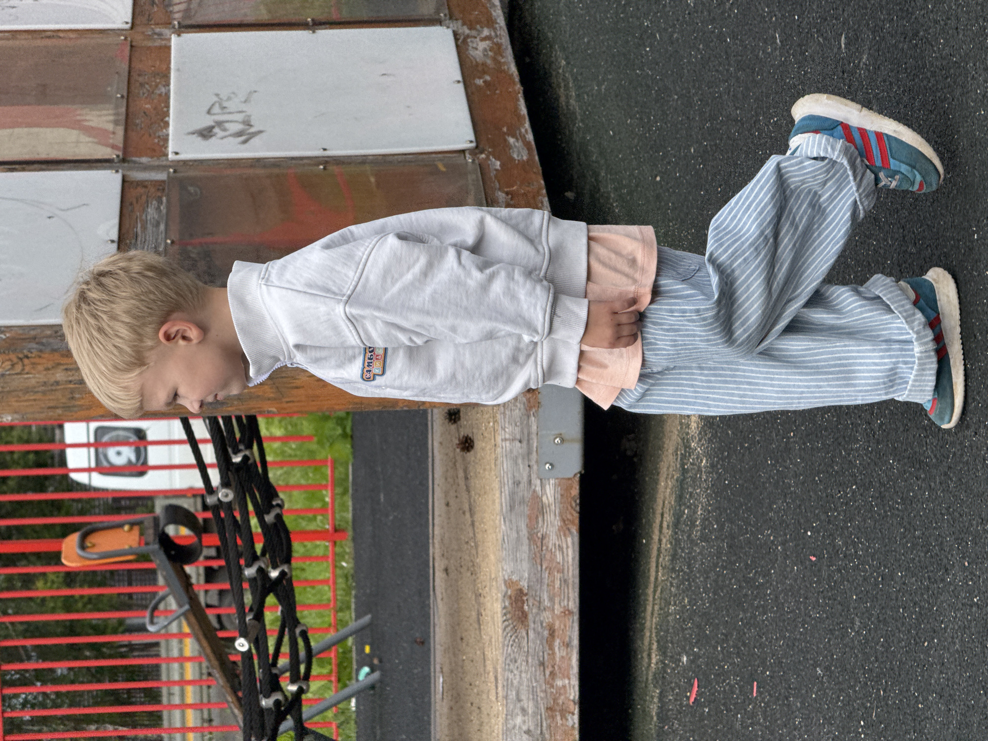 A young boy with blonde hair, wearing a light grey zippered sweatshirt over a peach t-shirt and light blue and white striped pants, walks to the right. He is wearing blue and red sneakers. The boy is on a playground with dark grey asphalt covering the ground. In the background, there is a wooden playground structure with some metal elements and what appears to be a red fence. Pinecones are visible on the wooden surface of the playground. The location is Bylovo, Russia.