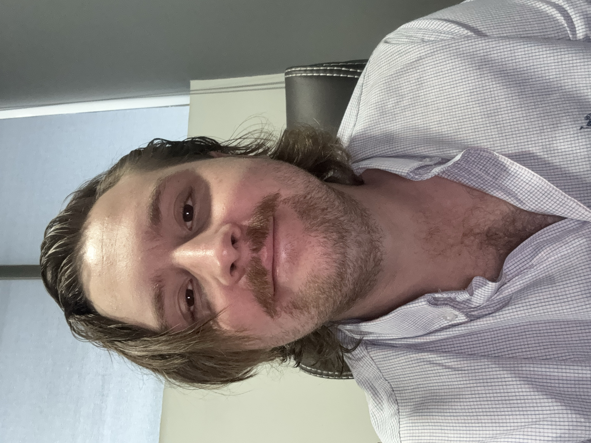 A close-up, indoor portrait depicts a man with light brown hair, a mustache, and a sparse beard. He is wearing a light-colored, checkered, button-down shirt. The background includes a light-colored wall and the dark back of a chair with white stitching. The image orientation suggests the photo was taken at an angle, possibly as a selfie. There is no information in the image that can confirm its location in Cuautitlán Izcalli, Mexico.