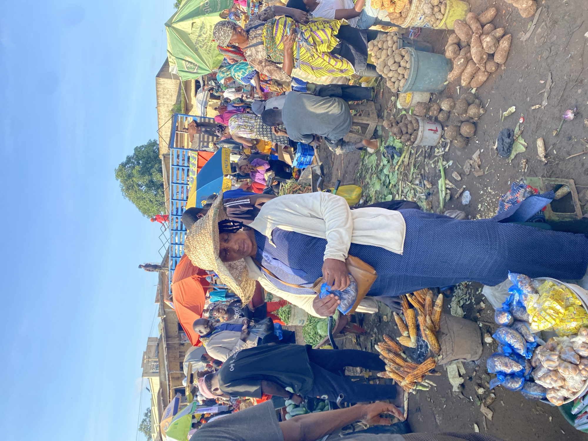 In Gura Top, Nigeria, this photograph captures a bustling outdoor market under a clear blue sky. A woman wearing a wide-brimmed straw hat and a blue dress stands center, holding a brown purse and a blue plastic bag. She appears to be examining the contents of the bag. Surrounding her are numerous market stalls and vendors selling a variety of produce. In the foreground, buckets overflow with corn on the cob, and a display of root vegetables, including potatoes and what appear to be yams, are piled on the ground. Behind the woman, other market-goers navigate the crowded space. In the background, several vehicles, including a truck, are visible, along with various market structures and awnings. The ground is a mix of dirt and scattered produce, indicating a busy and active marketplace.