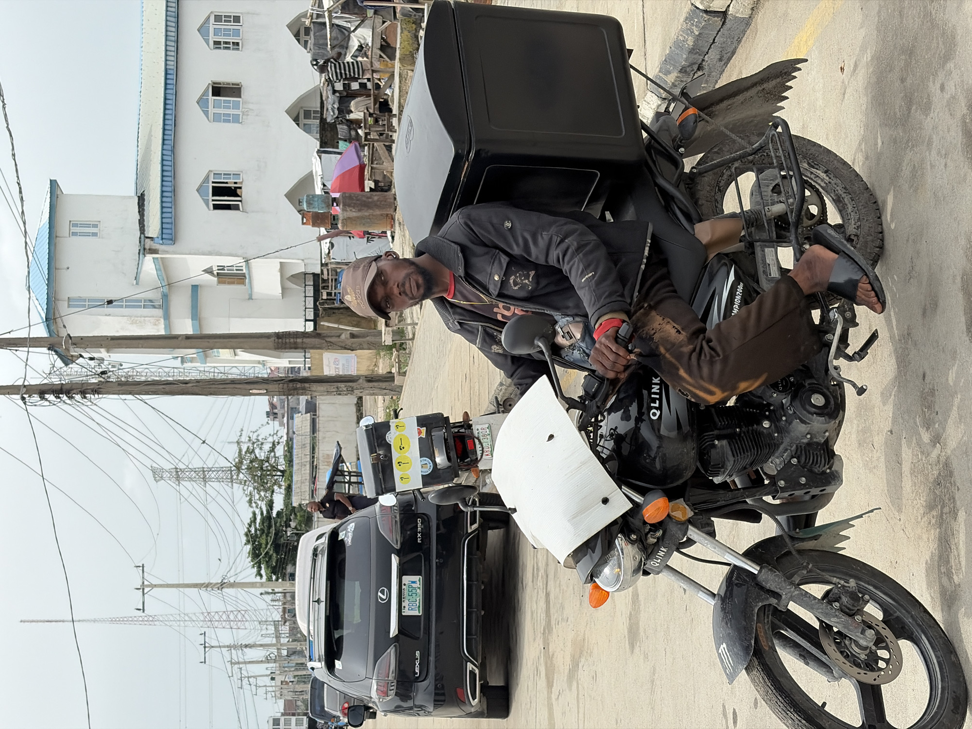 A man sits atop a black Qlink motorcycle with a large black delivery box mounted on the back in Igboefon, Nigeria. He wears a dark jacket, brown cap, and brown trousers and is looking at the camera. A black Lexus RX350 with license plate RBC56PY is parked behind the motorcycle. A building with white walls and blue accents is visible in the background. Above the car, overhead power lines are visible. The ground is paved.