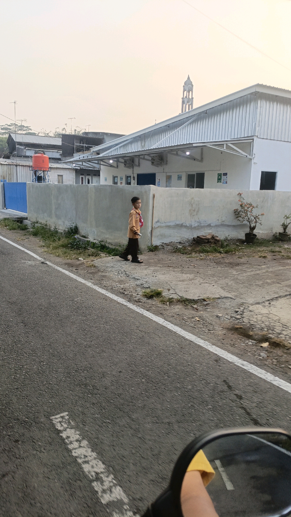 A young boy in an orange uniform with a red scarf walks along a concrete path beside a grey concrete wall. The wall is next to a grey asphalt road with a white painted line. In the background, a white building with a metal roof has a mosque minaret visible beyond it. An orange water tank sits atop a small structure on the left. The scene appears to be in a suburban or industrial area of Salatiga, Indonesia, possibly near a school given the boy's uniform. The lighting suggests it is either early morning or late afternoon.