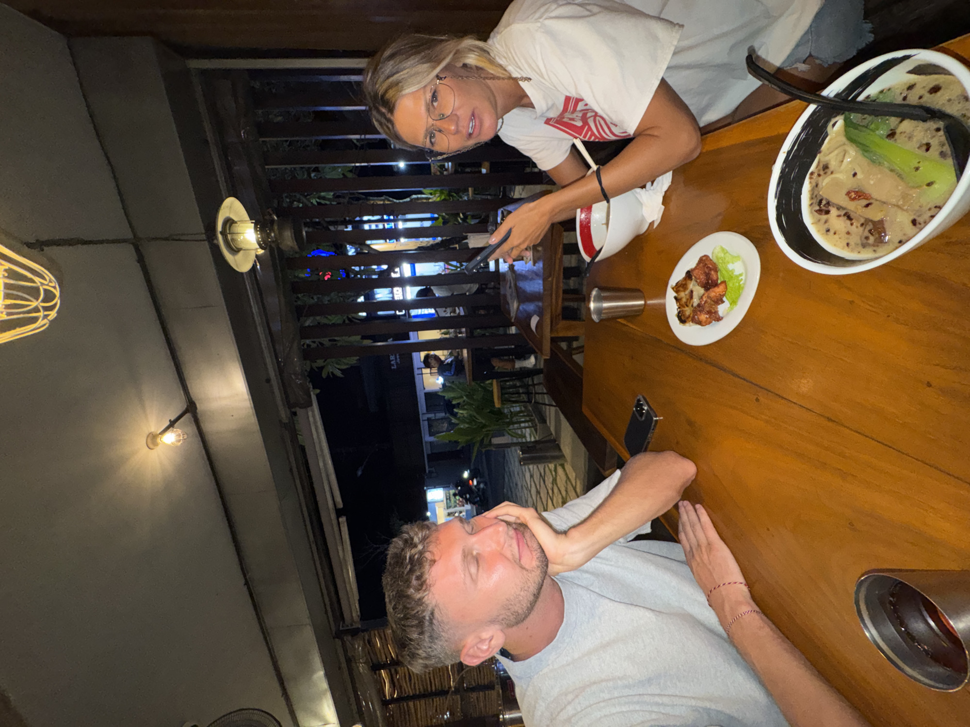 The image shows two individuals seated at a wooden table in what appears to be an outdoor dining setting in North Kuta, Indonesia. A woman with blonde hair and glasses is holding a phone. The man next to her is with closed eyes and resting his head in his hand. On the table is a bowl of soup, a plate with chicken, and various other items. The setting includes ambient lighting and foliage, suggesting an evening atmosphere. Outdoor structures and distant lights indicate a city environment.