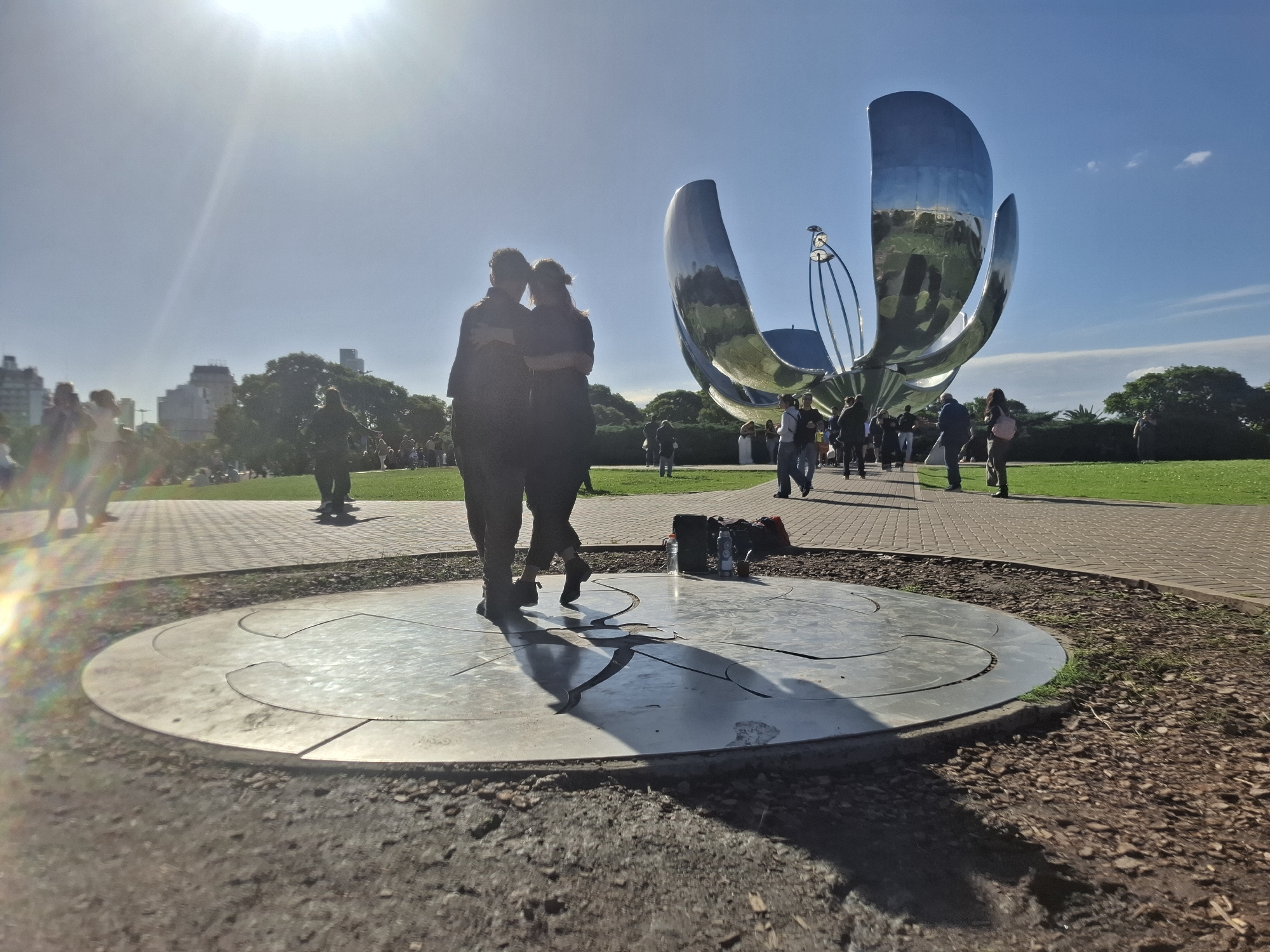 A street view shot in Buenos Aires, Argentina, features a couple dancing the tango on a circular platform, located near the Floralis Generica, a large metallic flower sculpture. The sculpture stands behind the dancing couple, with several other people walking around in the background. The sky is blue, and the sun is shining brightly, creating shadows and casting light across the scene. The ground surrounding the platform consists of dirt and mulch, while a brick pathway stretches behind the sculpture. Other pedestrians and trees are scattered throughout the background. Buildings are visible in the far background. A bag and water bottle are placed near the platform.
