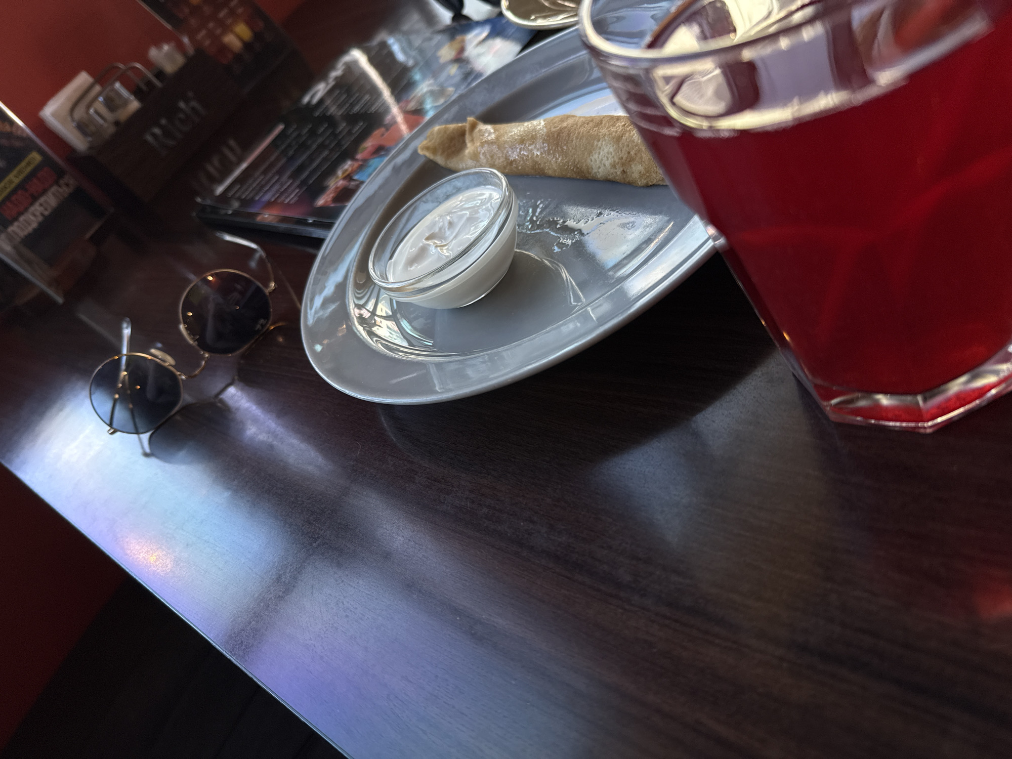 The image captures a top-down view of a table in what appears to be a cafe or restaurant, focusing on a meal and other items. The dominant subject is a gray oval plate holding a rolled pancake or crepe, dusted lightly with powdered sugar. Next to the pancake is a small clear glass bowl filled with a white creamy sauce, likely sour cream or a similar condiment. To the right of the plate, a clear glass contains a vibrant red beverage, possibly juice or a berry drink.
Further to the left on the dark wooden table, a pair of dark-lensed sunglasses with gold frames rests. In the blurred background, several objects are visible, including a menu holder with the word "Rich" clearly legible on it, and what looks like another menu or magazine. The table surface reflects some light, creating subtle highlights. The overall impression is of a casual dining setting, with someone enjoying a sweet dish and a drink. The lighting suggests an indoor environment, possibly during the daytime.